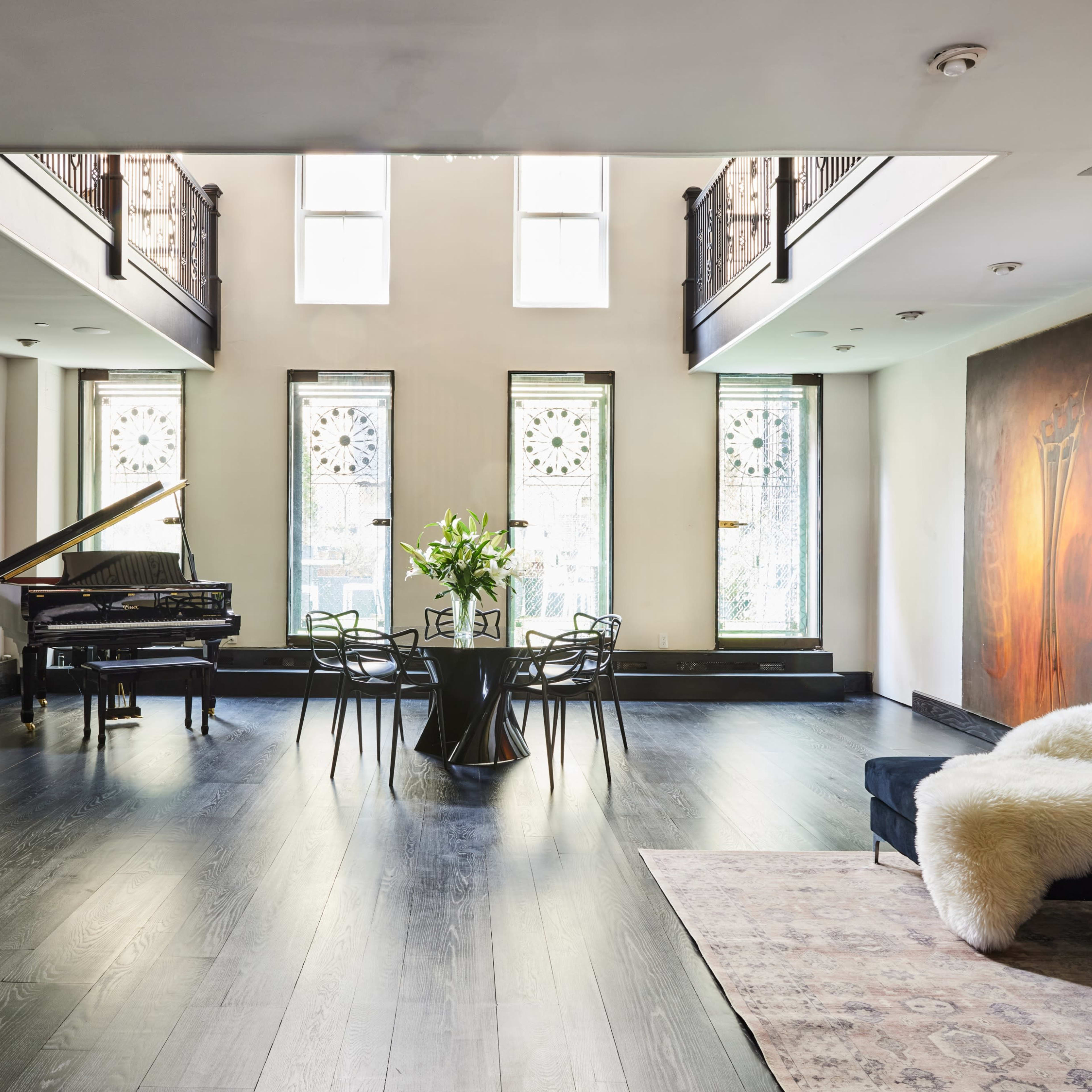 The spacious room features a grand piano, a circular dining table with chairs, and large windows with decorative patterns, along with artwork on the walls.