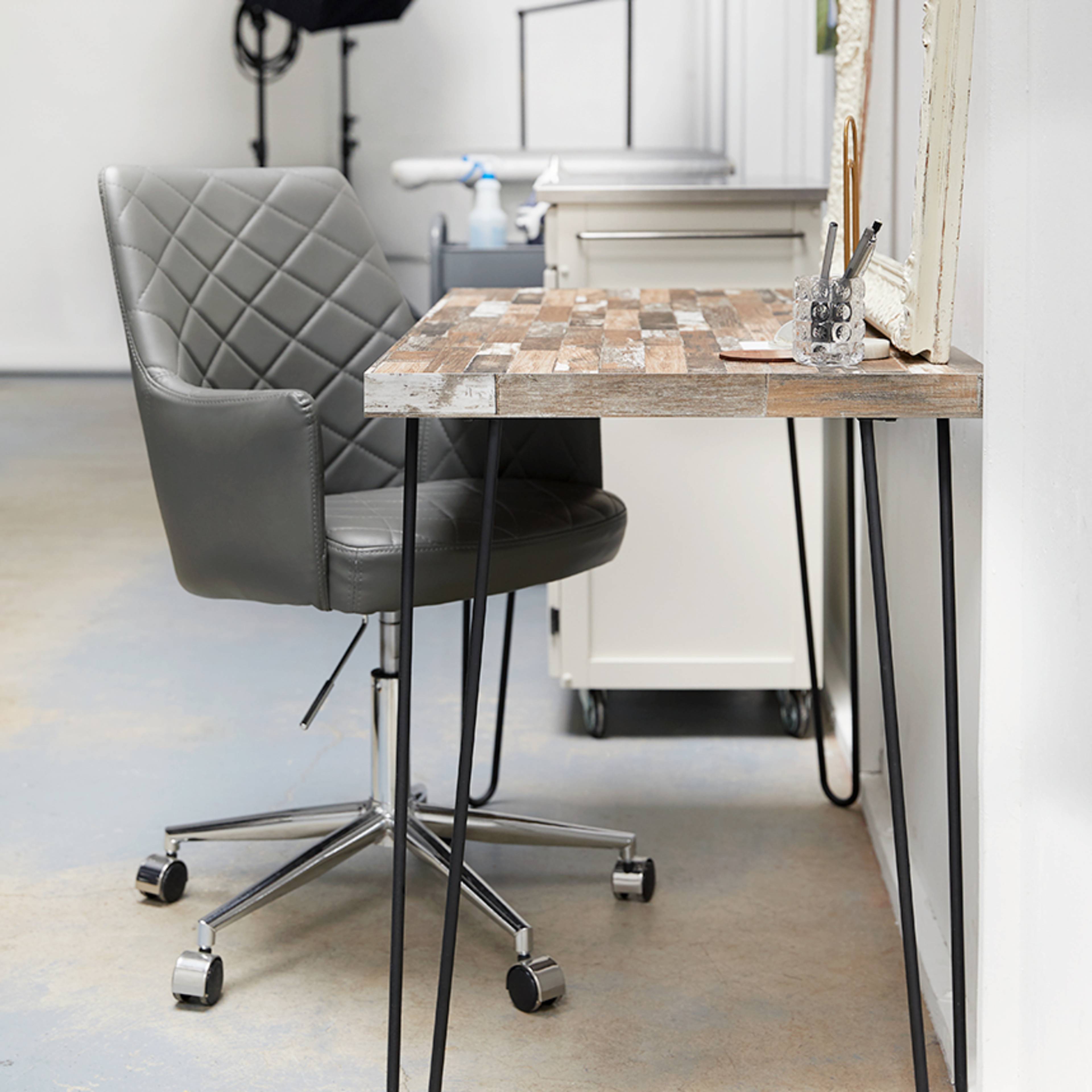 A modern workspace features a gray swivel chair next to a wooden desk with metal legs, along with a framed mirror and a photography umbrella in the background.