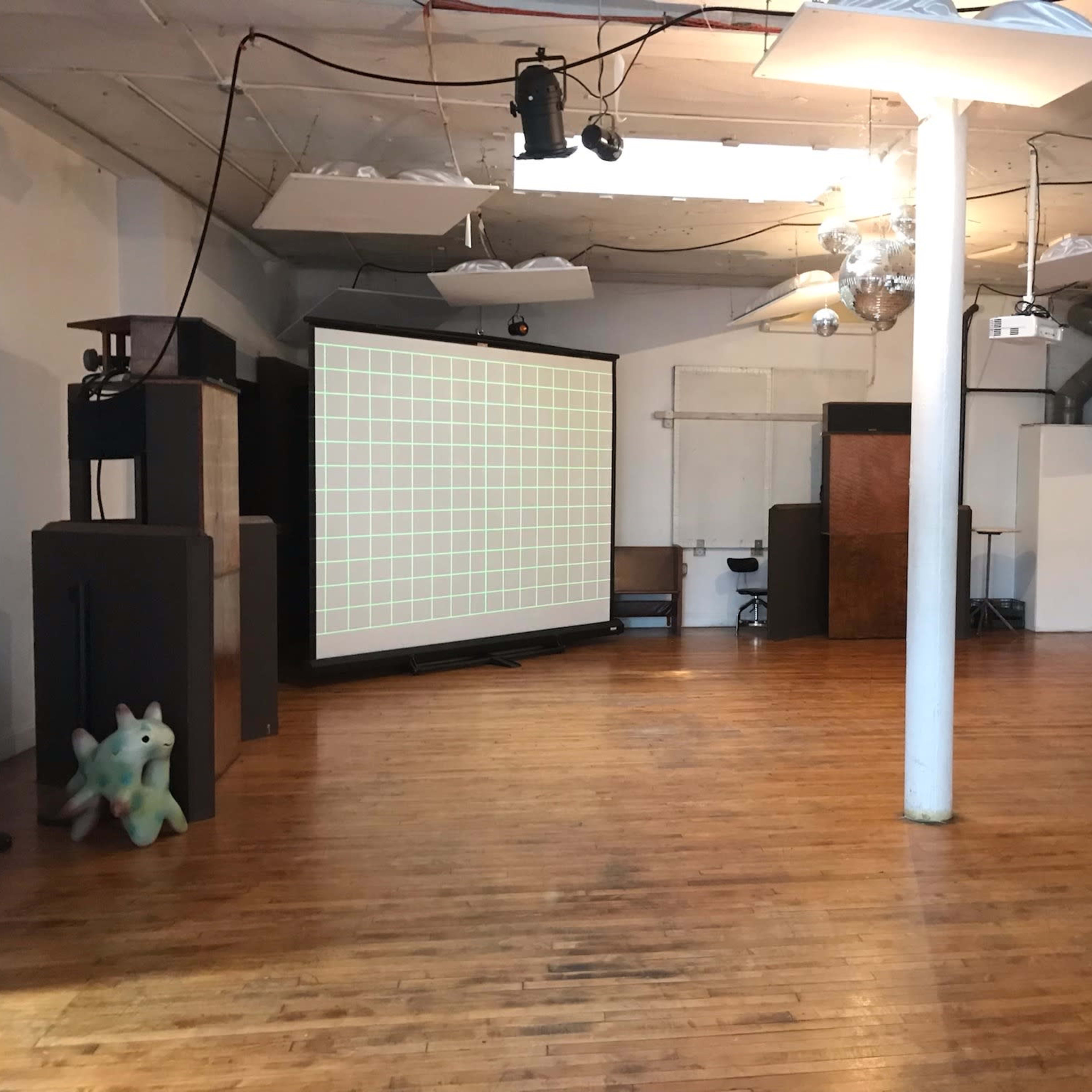 An empty indoor space with wooden flooring, featuring a large projection screen, audio equipment, and a colorful stuffed figure in the corner.