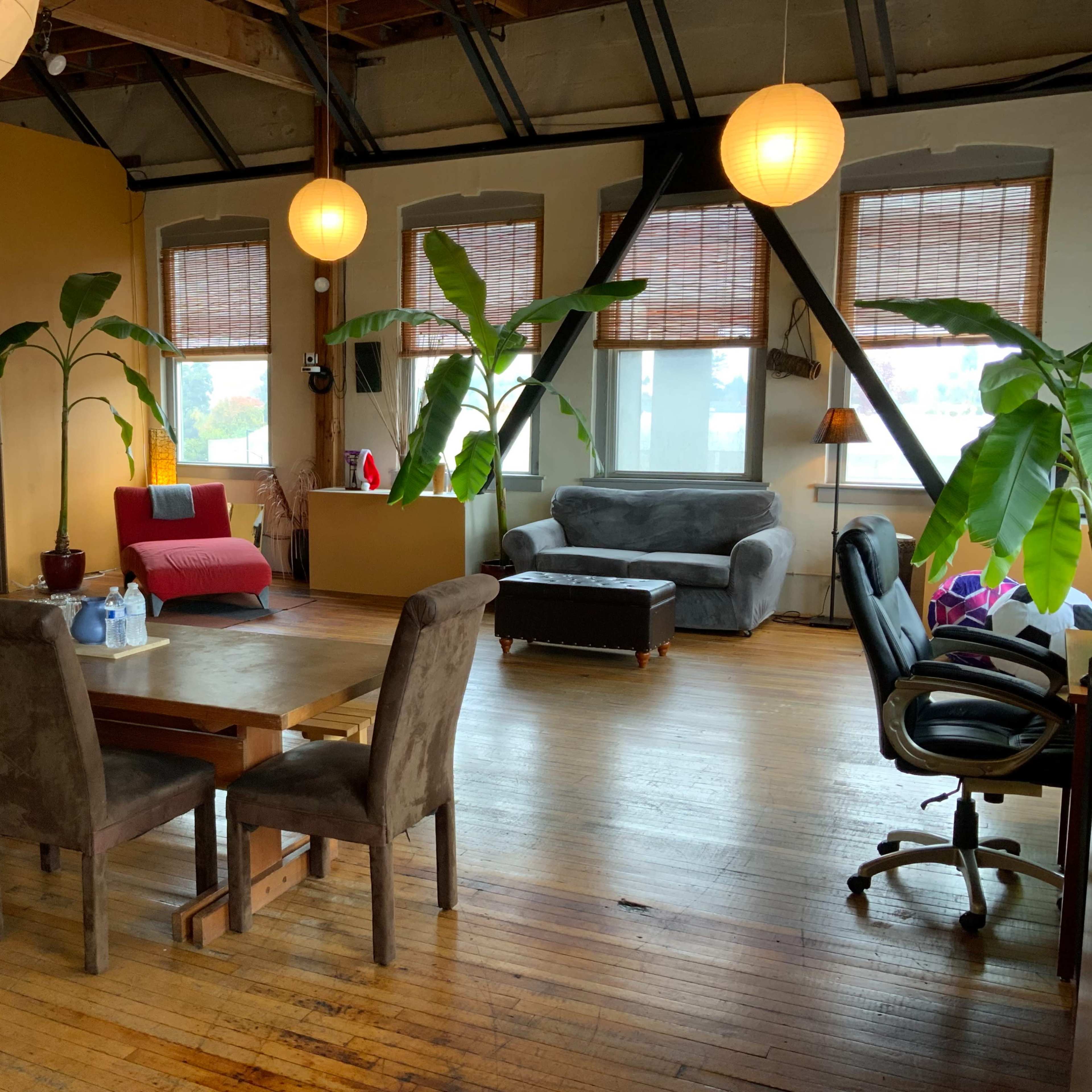 A spacious, sunlit loft with a dining area featuring a wooden table and chairs, alongside a living area with various seating options and large windows adorned with potted plants.