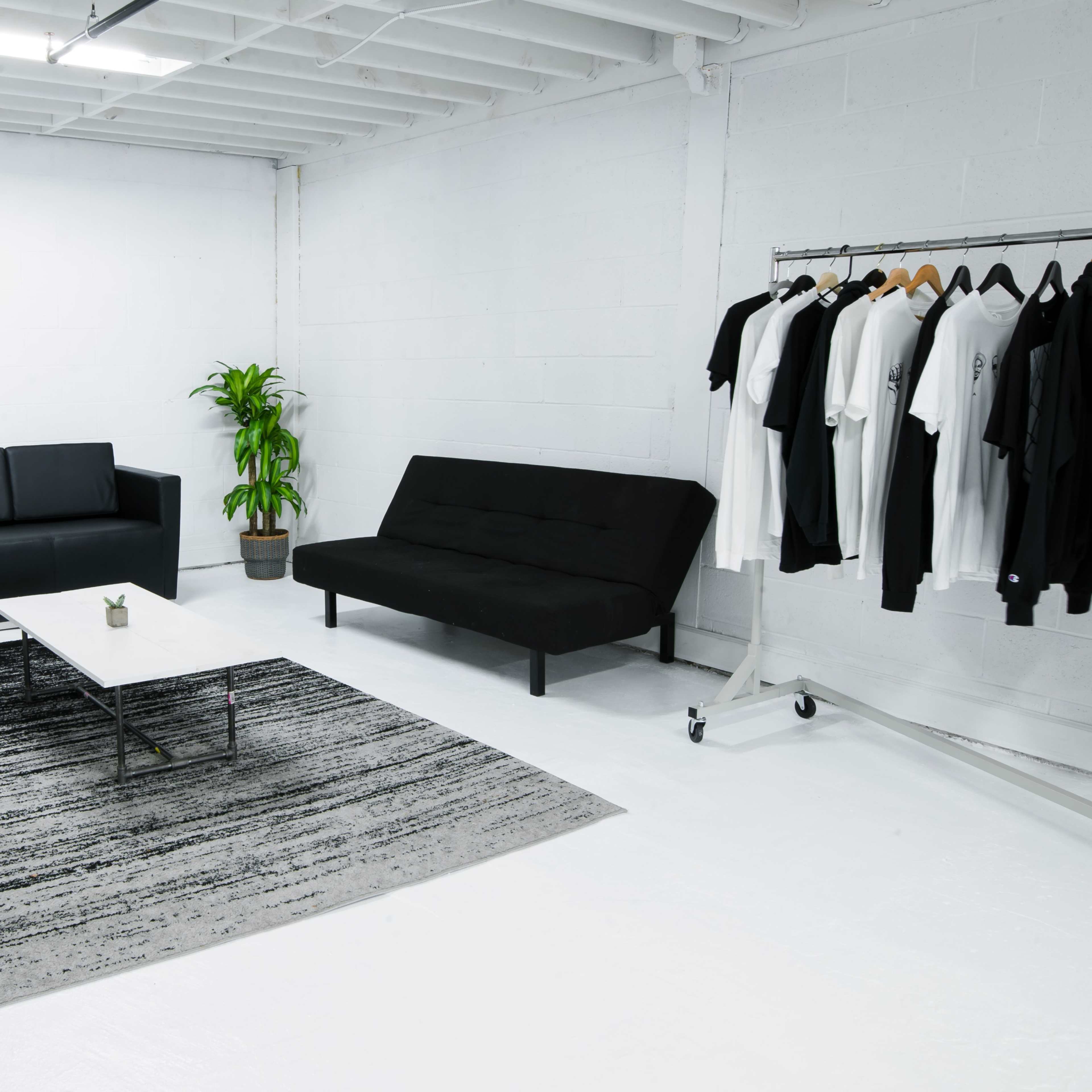 A minimalist white room featuring two sofas, a coffee table, a plant, a rug, and a clothing rack with hanging garments.
