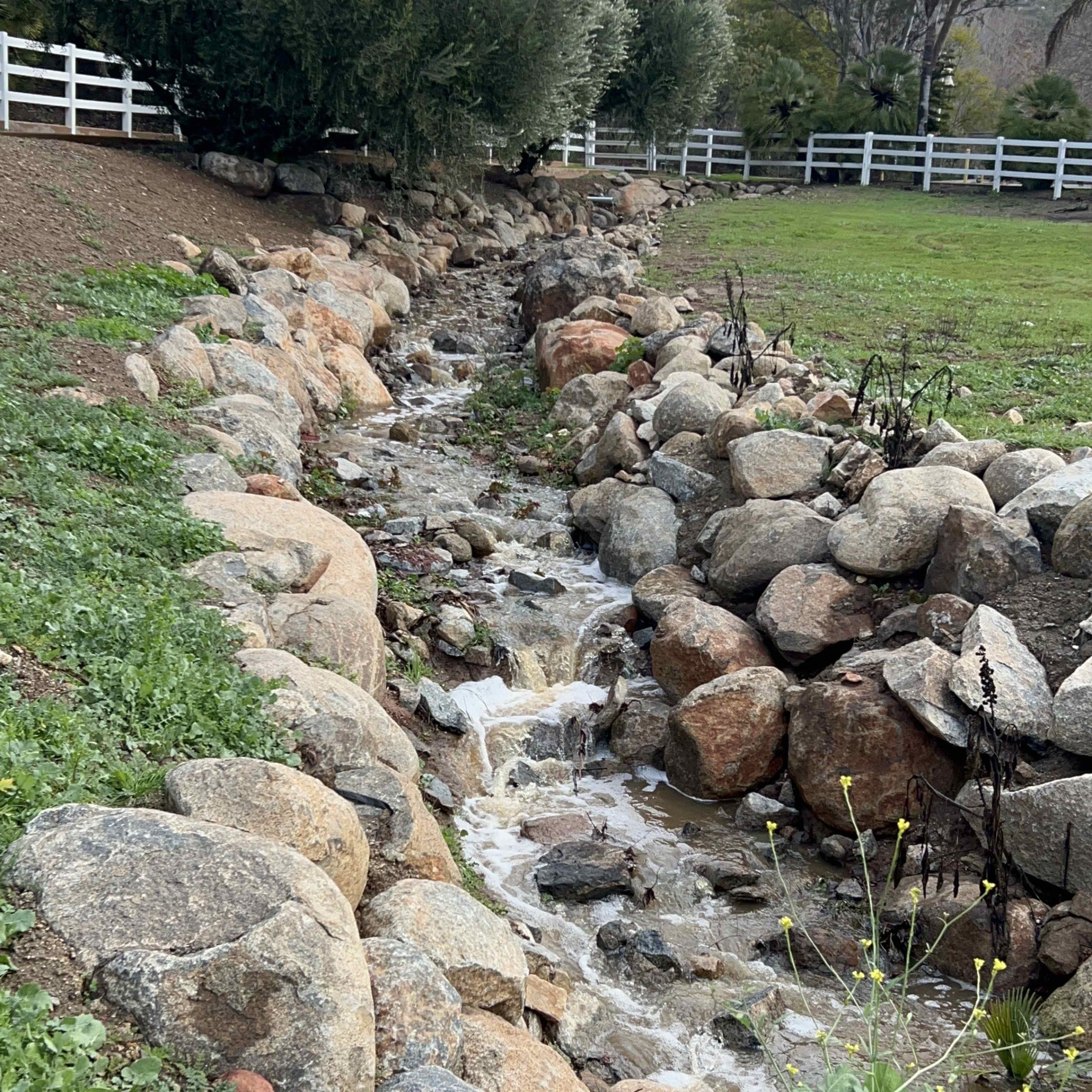A narrow water channel flows between large rocks in a landscaped area with green grass and shrubs.