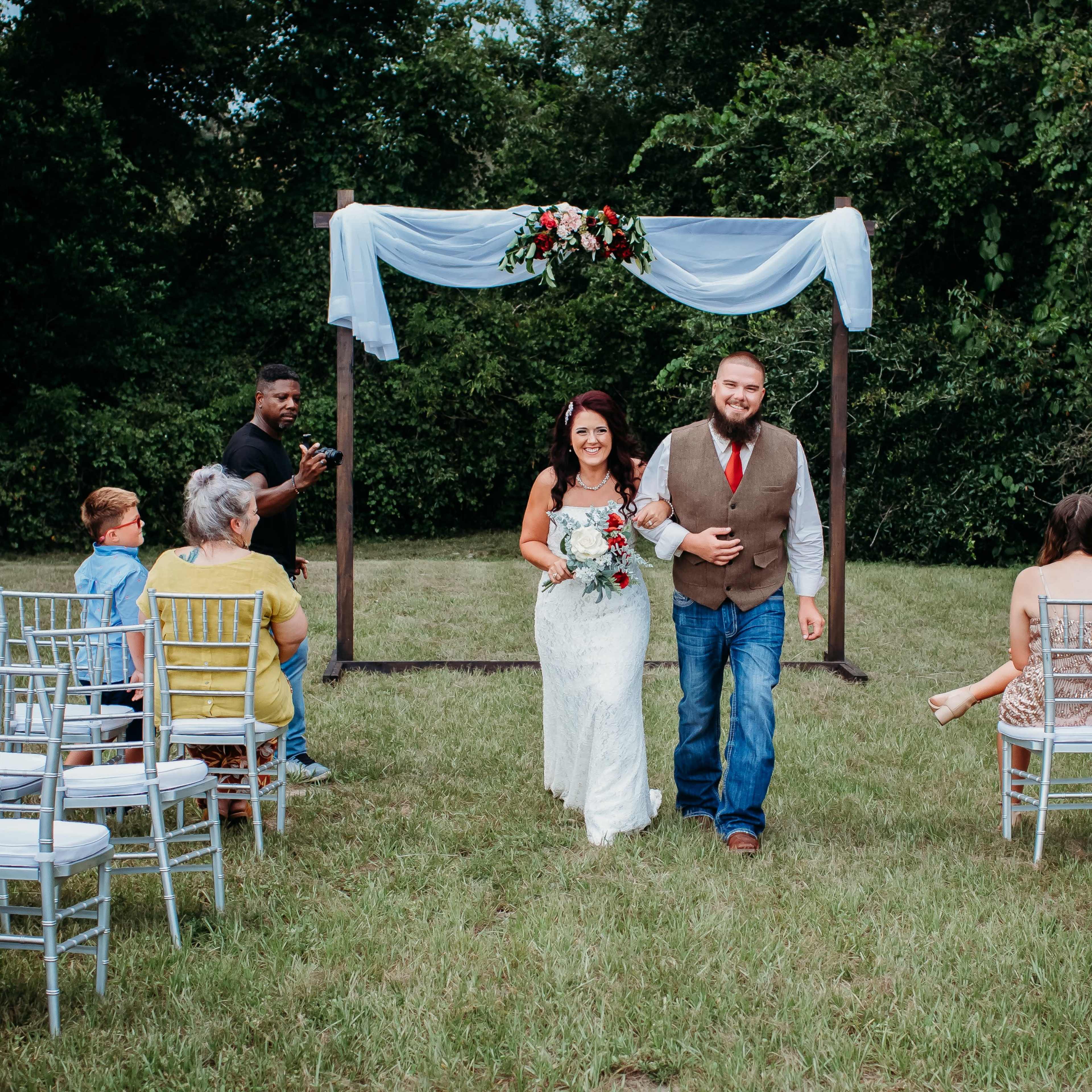 A bride and groom walk down an aisle together under an arch adorned with flowers, while seated guests look on in a grassy outdoor setting.