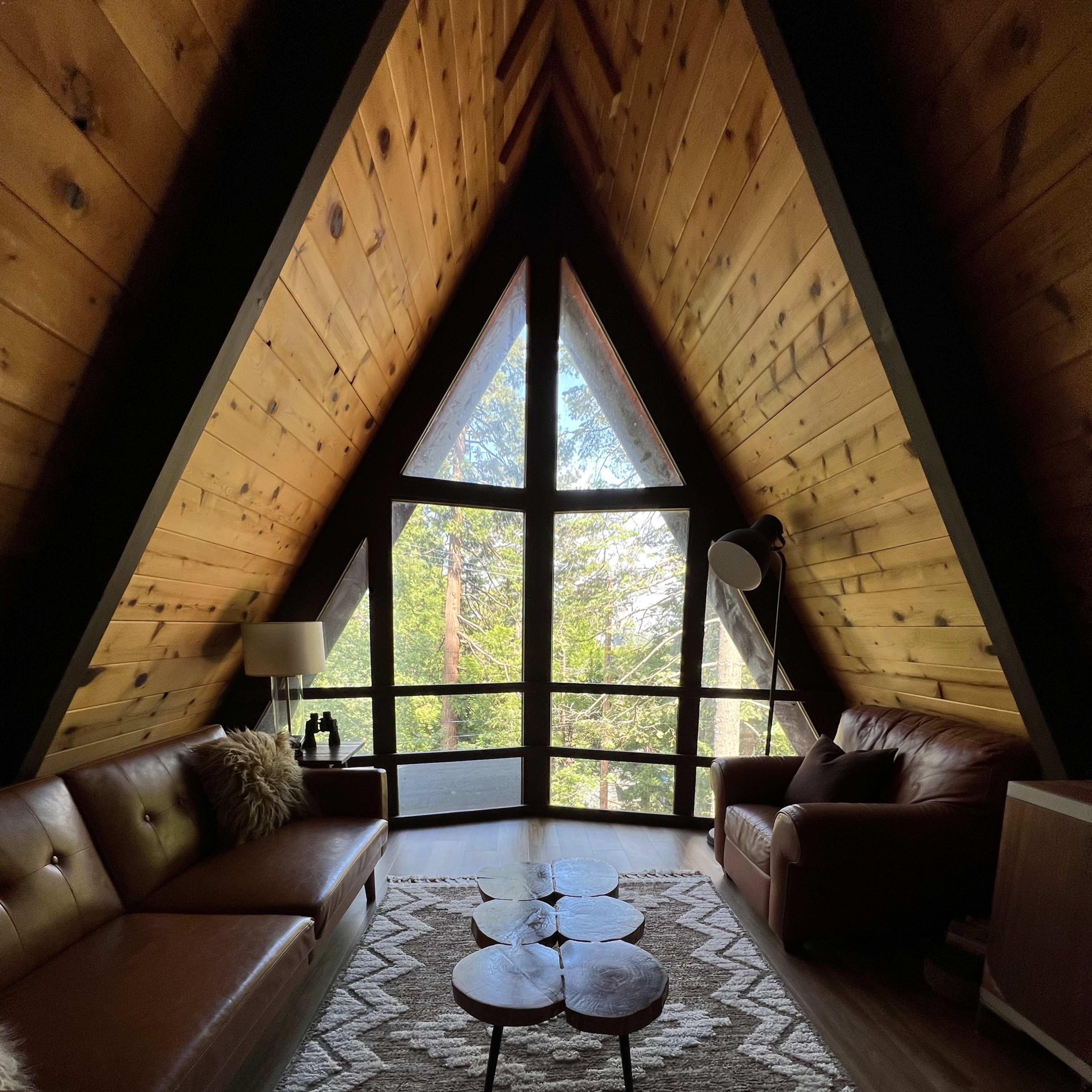 The interior of an A-frame cabin features wooden walls, a large triangular window, two leather sofas, a patterned rug, and a coffee table made of three wooden slabs.