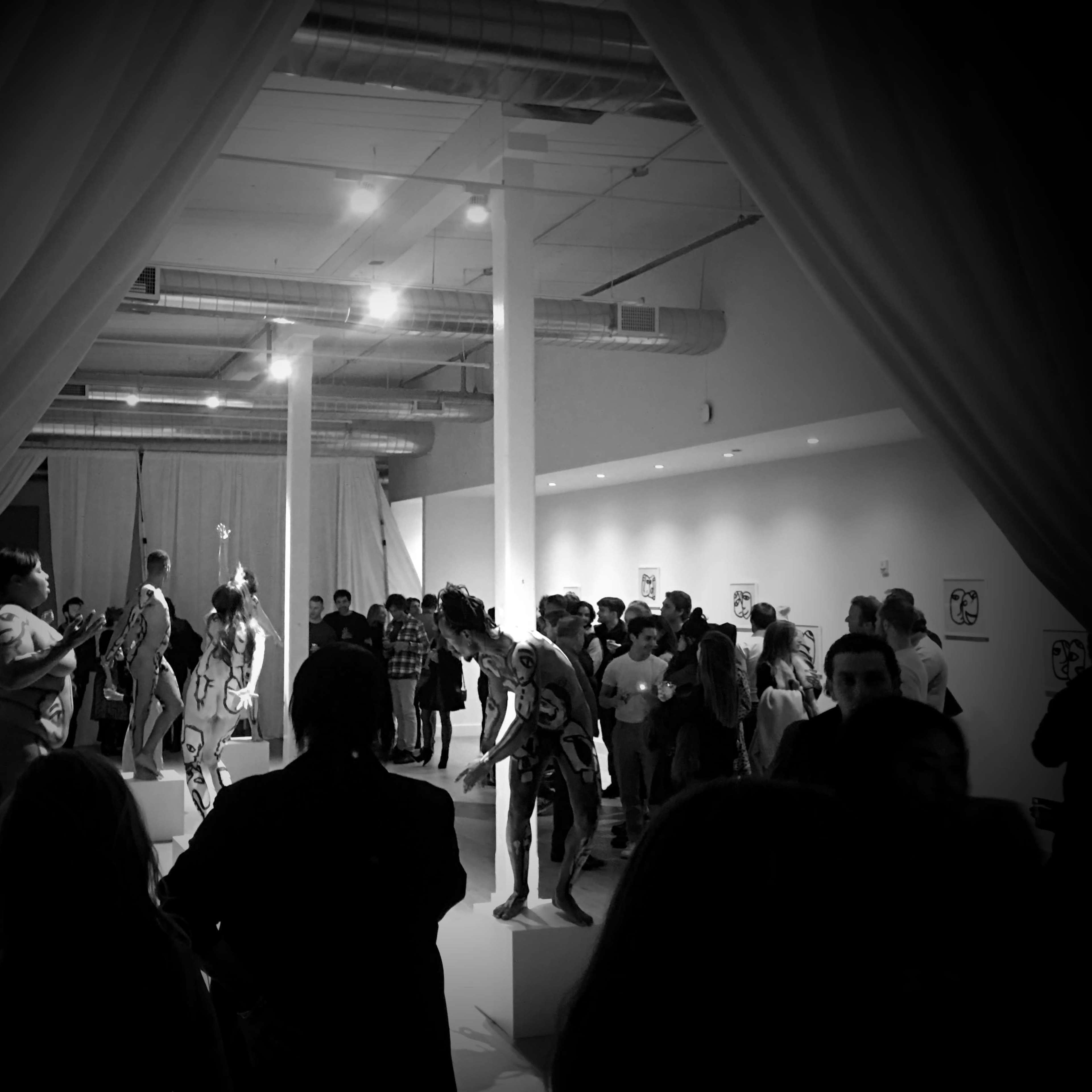 A crowd attends an art exhibit featuring performers in body paint amidst a white-walled gallery space.