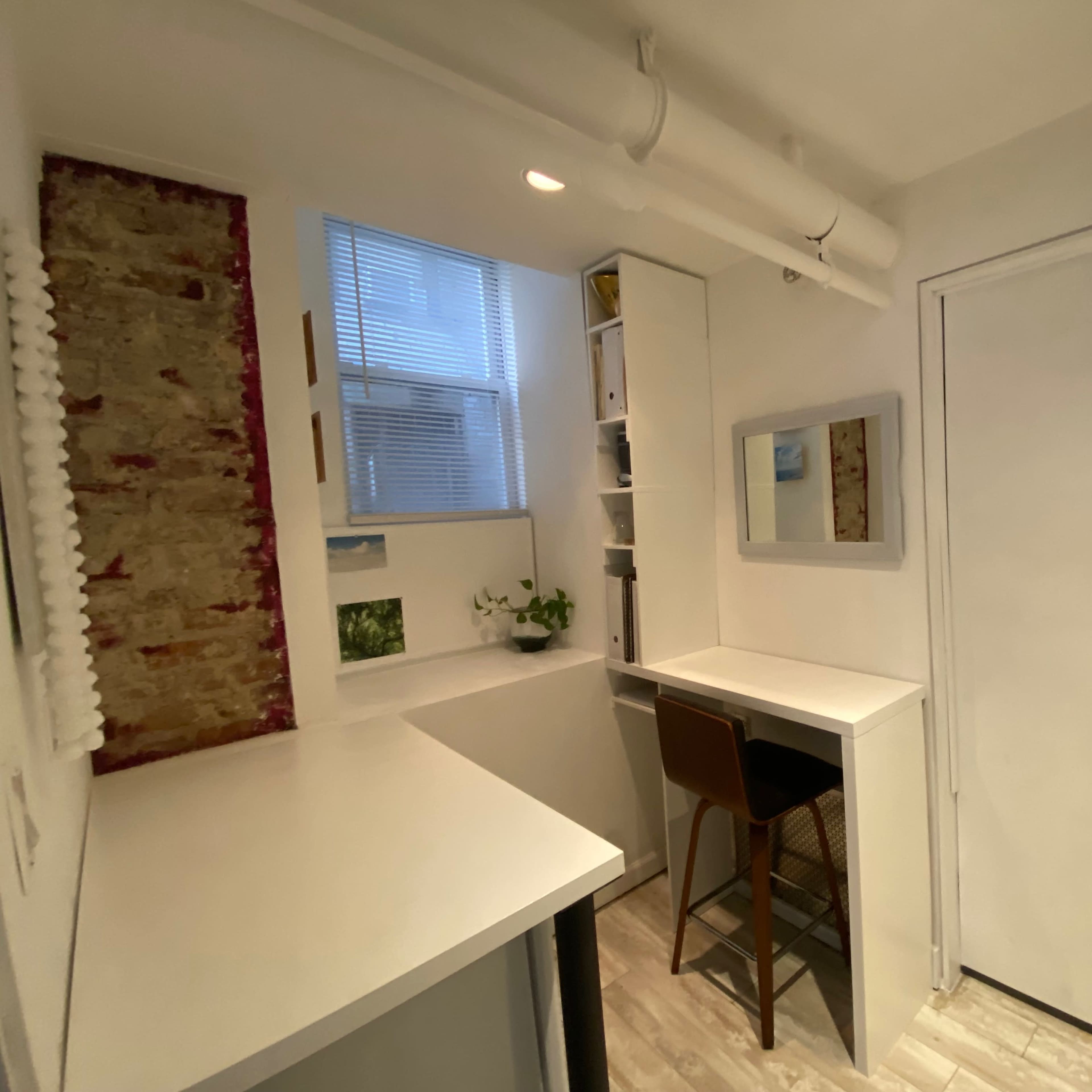 A small, minimalist workspace with a desk and a chair, a mirror on the wall, and a window allowing natural light to enter.