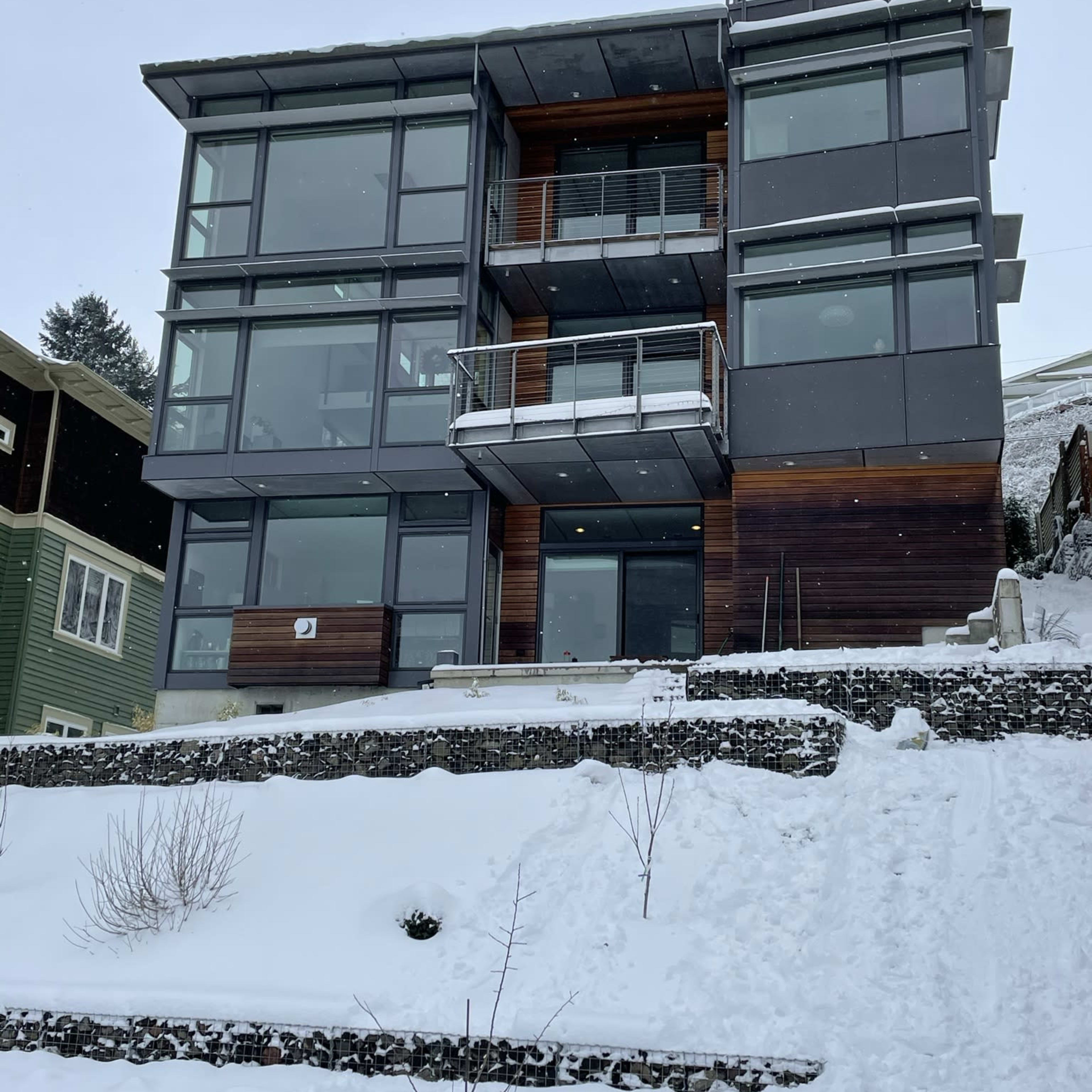 A modern, multi-story house with large glass windows and wooden accents is surrounded by snow and slopes.