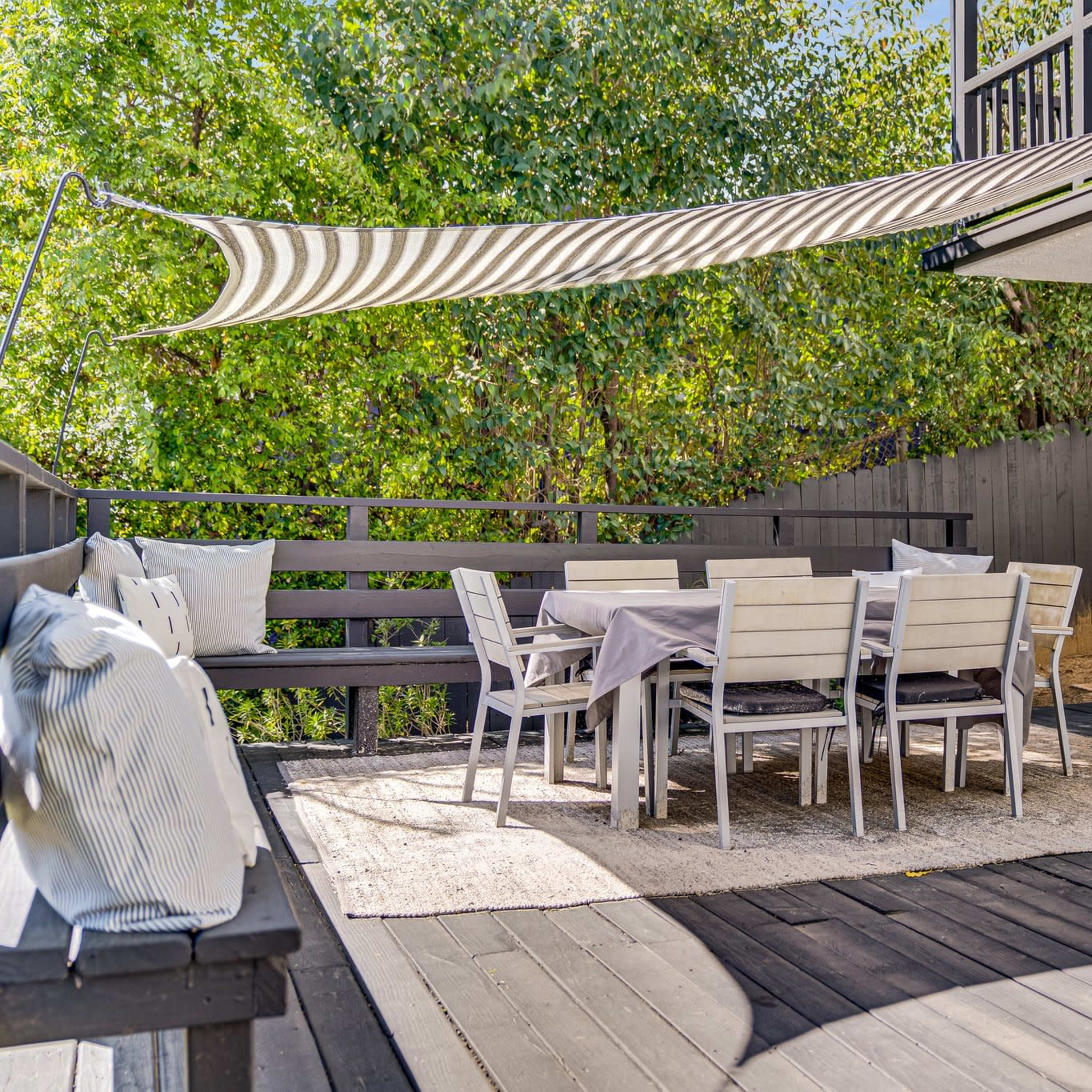 A spacious wooden deck features a dining table surrounded by chairs, with a shaded area provided by a striped awning.