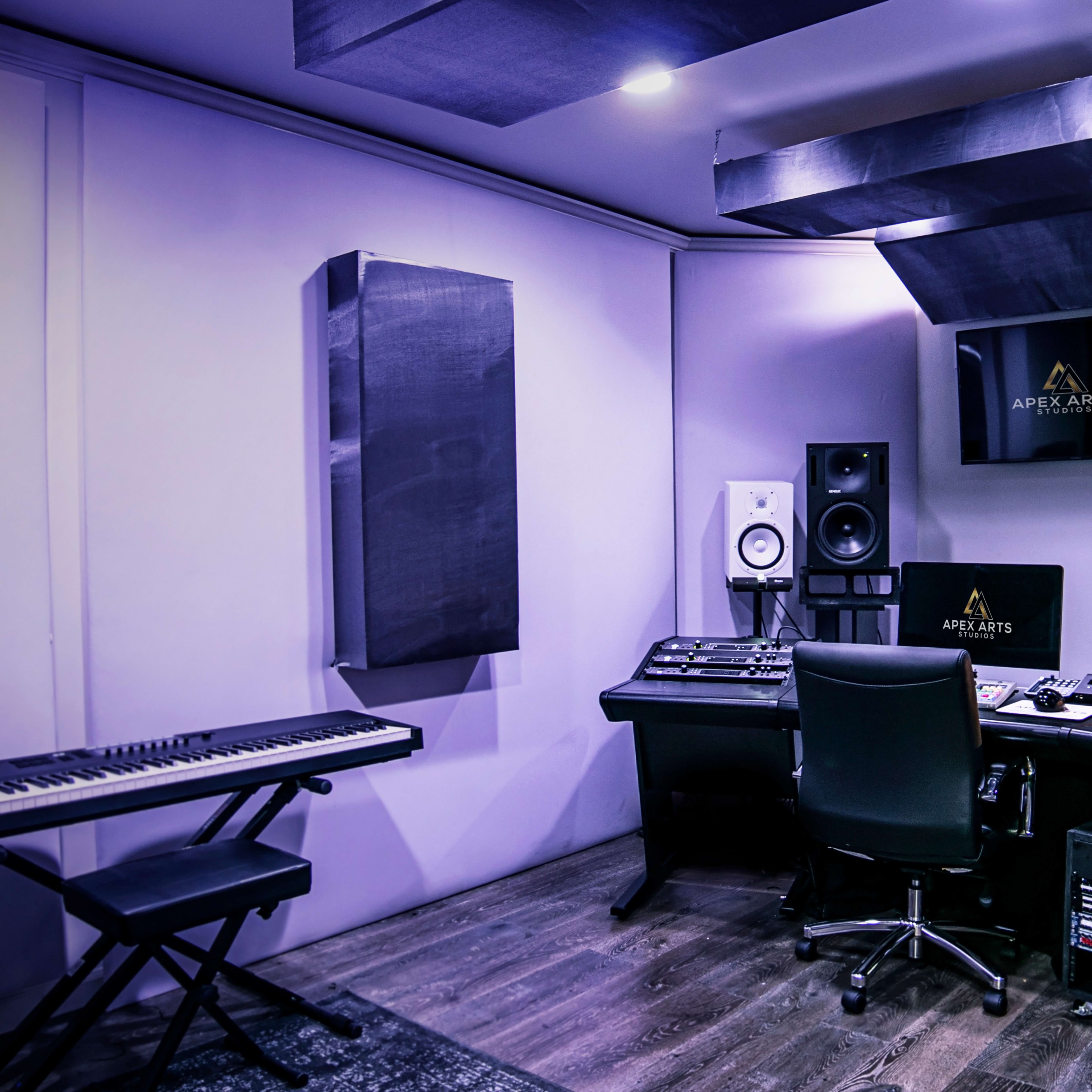 A music studio with a black desk, audio equipment, speakers, a keyboard, and acoustic treatment on the walls.