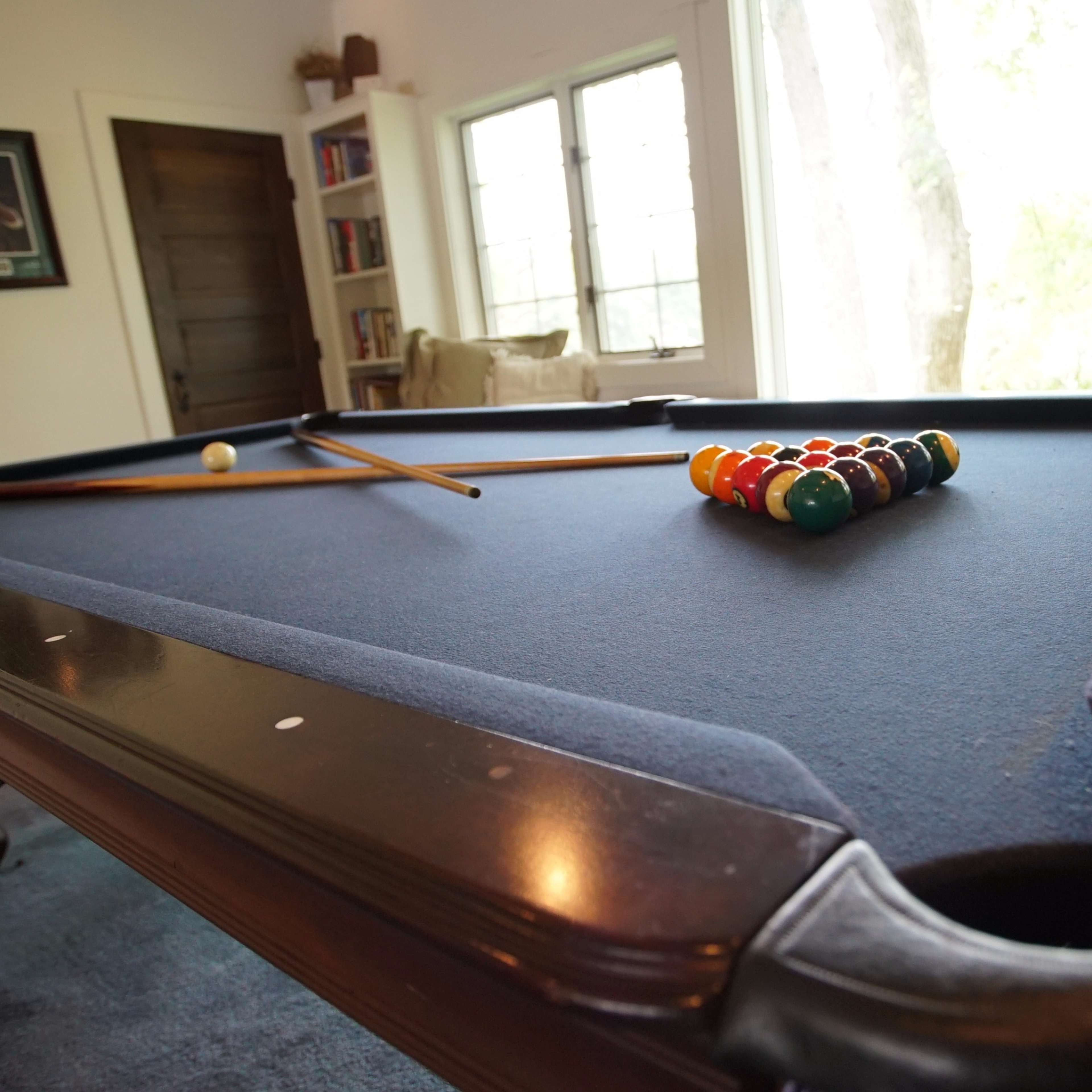 A pool table with a complete set of balls arranged in a triangle is positioned in a well-lit room featuring large windows.