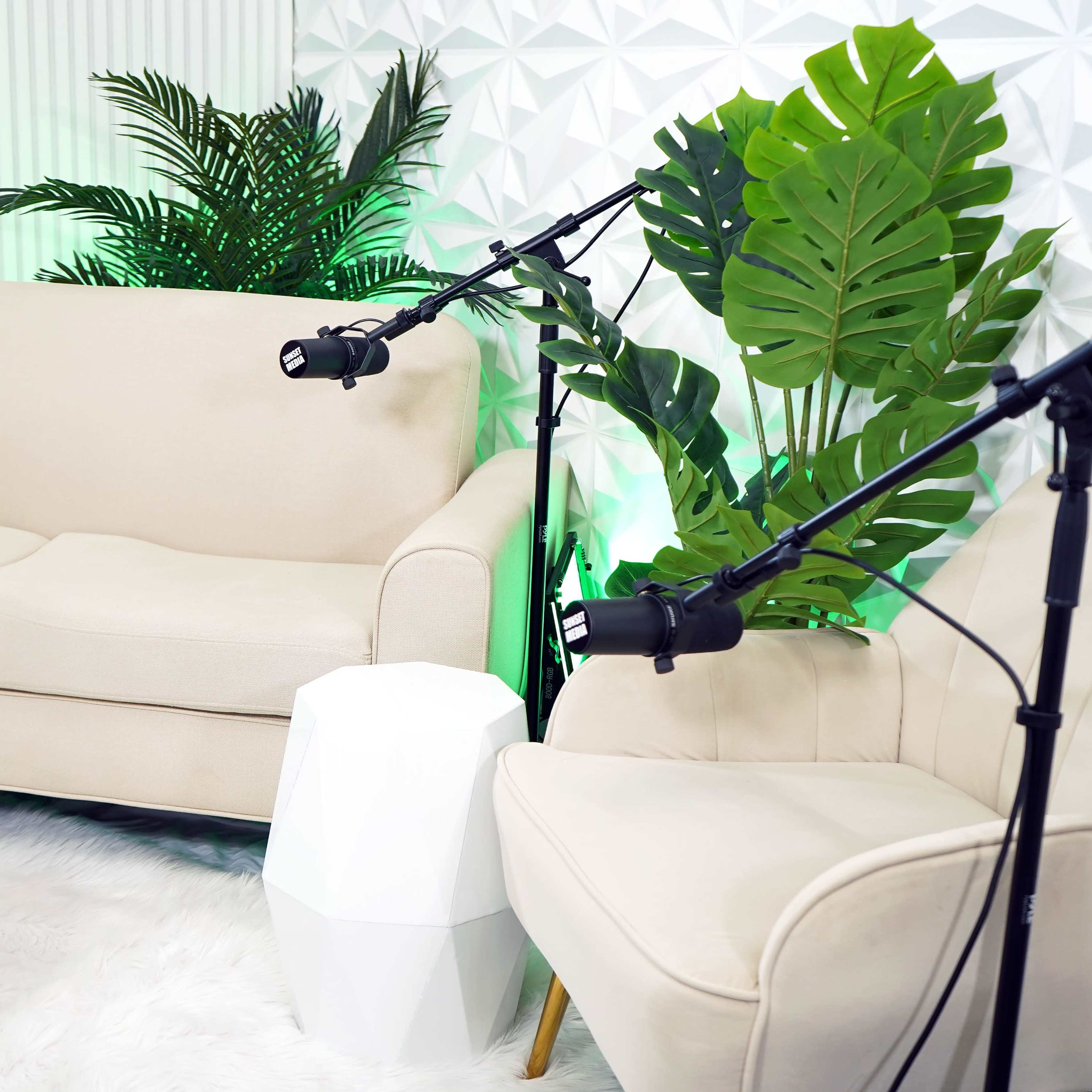 A modern seating area with a beige sofa, a white chair, and two adjustable lights, surrounded by large green plants against a textured wall.