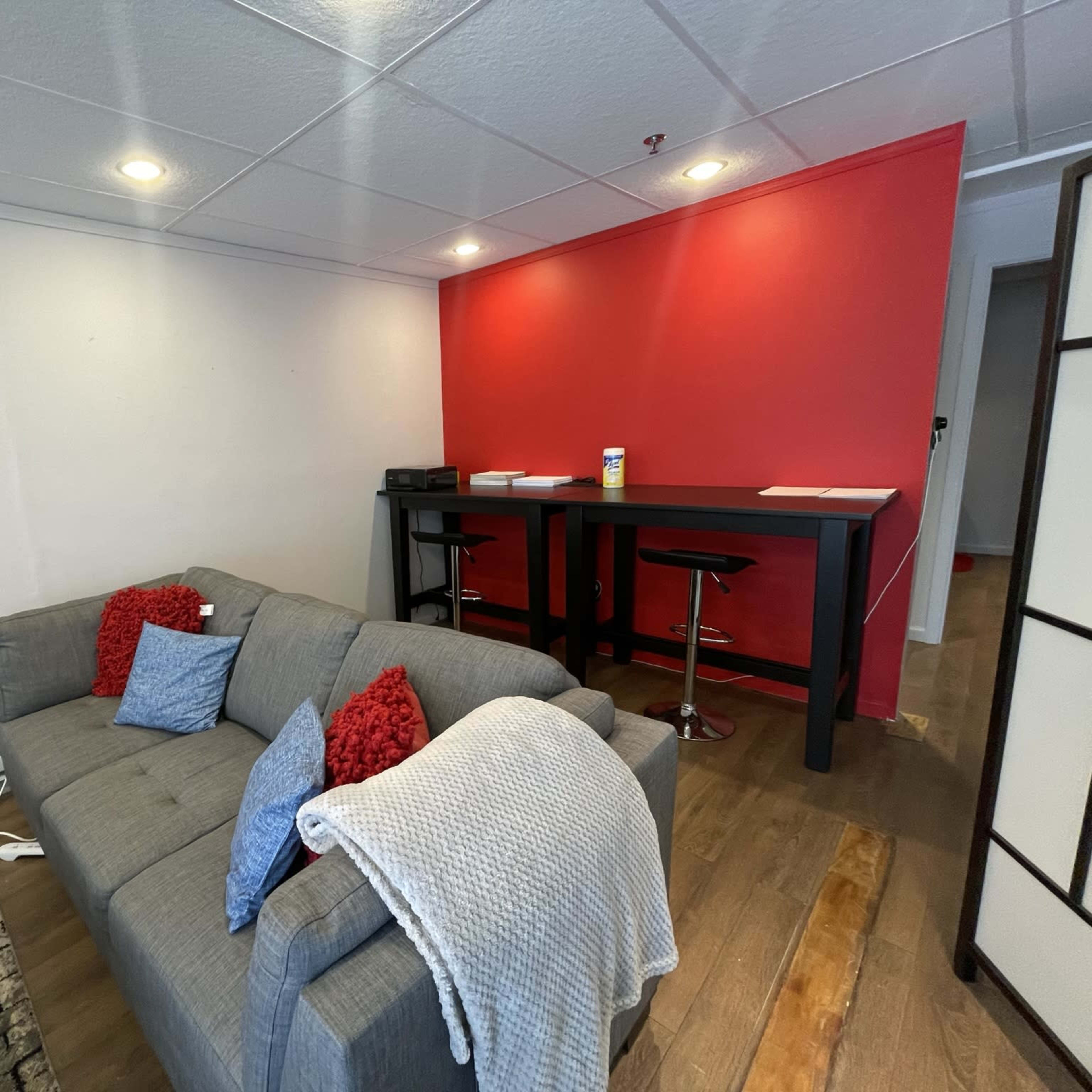 A gray couch with red pillows sits in a room next to a black bar table with two stools against a red wall.