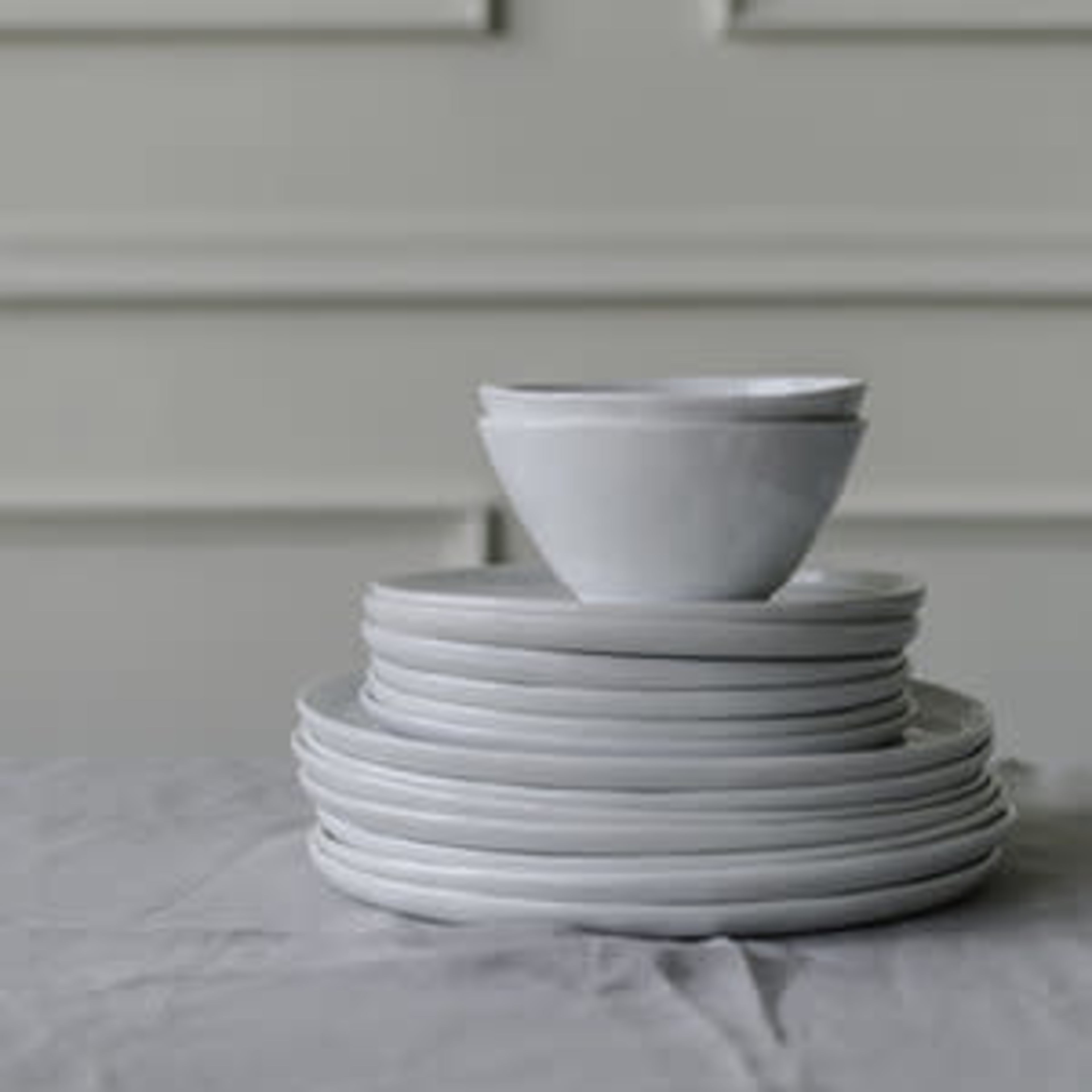 A stack of white plates topped with a single white bowl sits on a gray tablecloth in front of a light-colored wall.