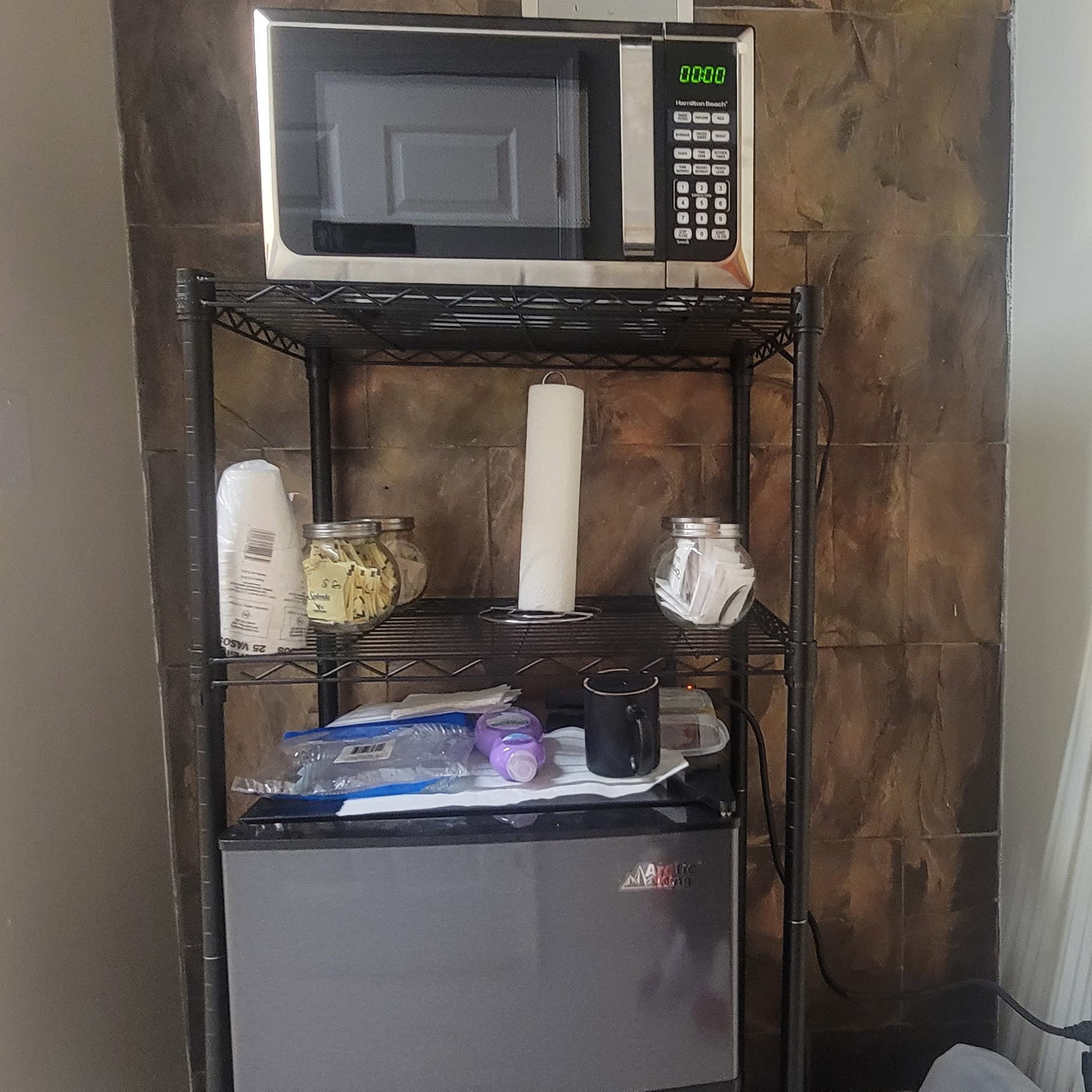 A microwave is placed on a metal shelf above a small refrigerator, surrounded by various kitchen items, against a wall with tiled decor.