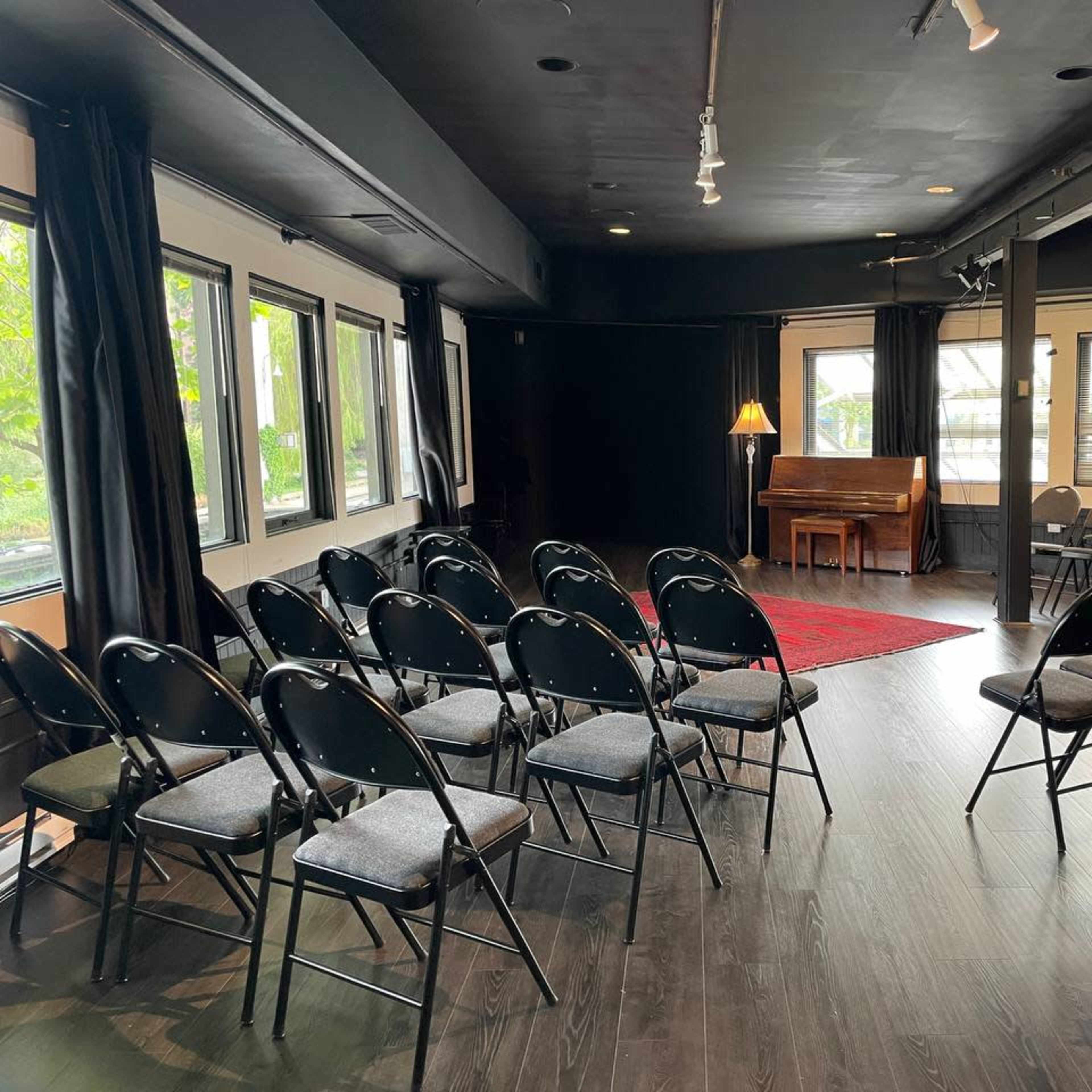 A small indoor performance space features rows of folding chairs facing a red rug and a piano, with large windows on one side.