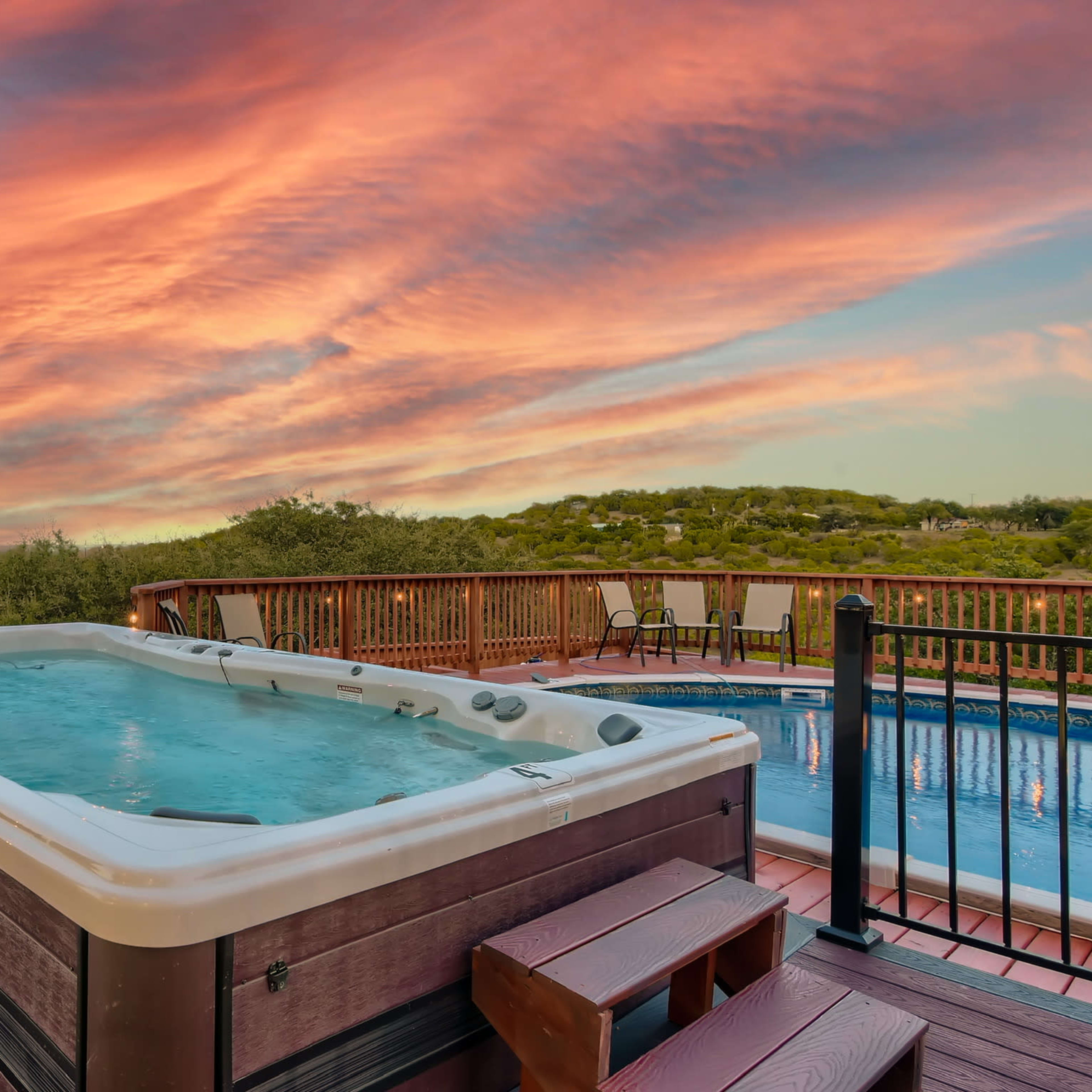 A hot tub is positioned on a wooden deck overlooking a pool, with a colorful sunset sky in the background.