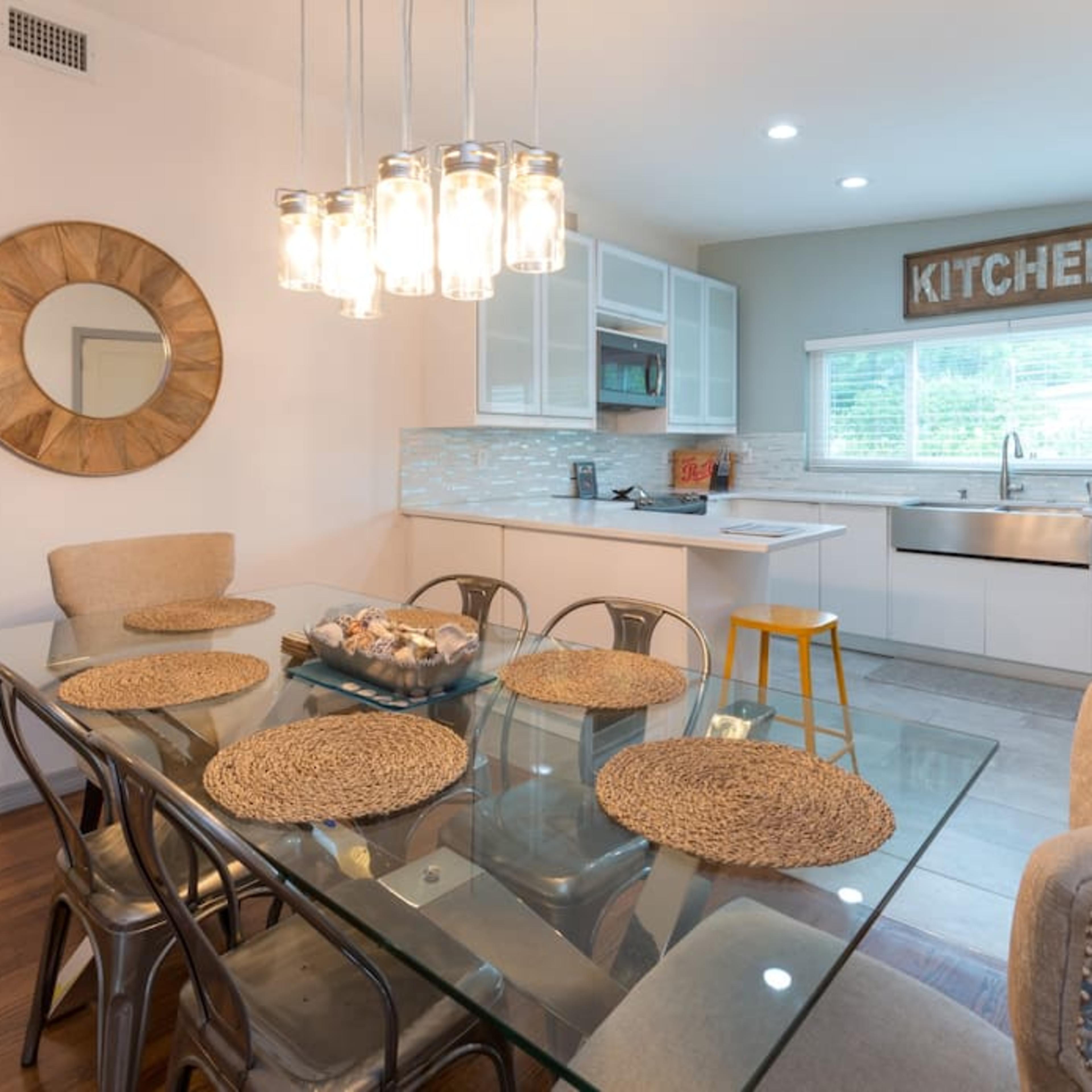 A modern dining area features a glass table with woven placemats, surrounded by upholstered chairs, overlooking a kitchen with white cabinetry and stainless steel appliances.