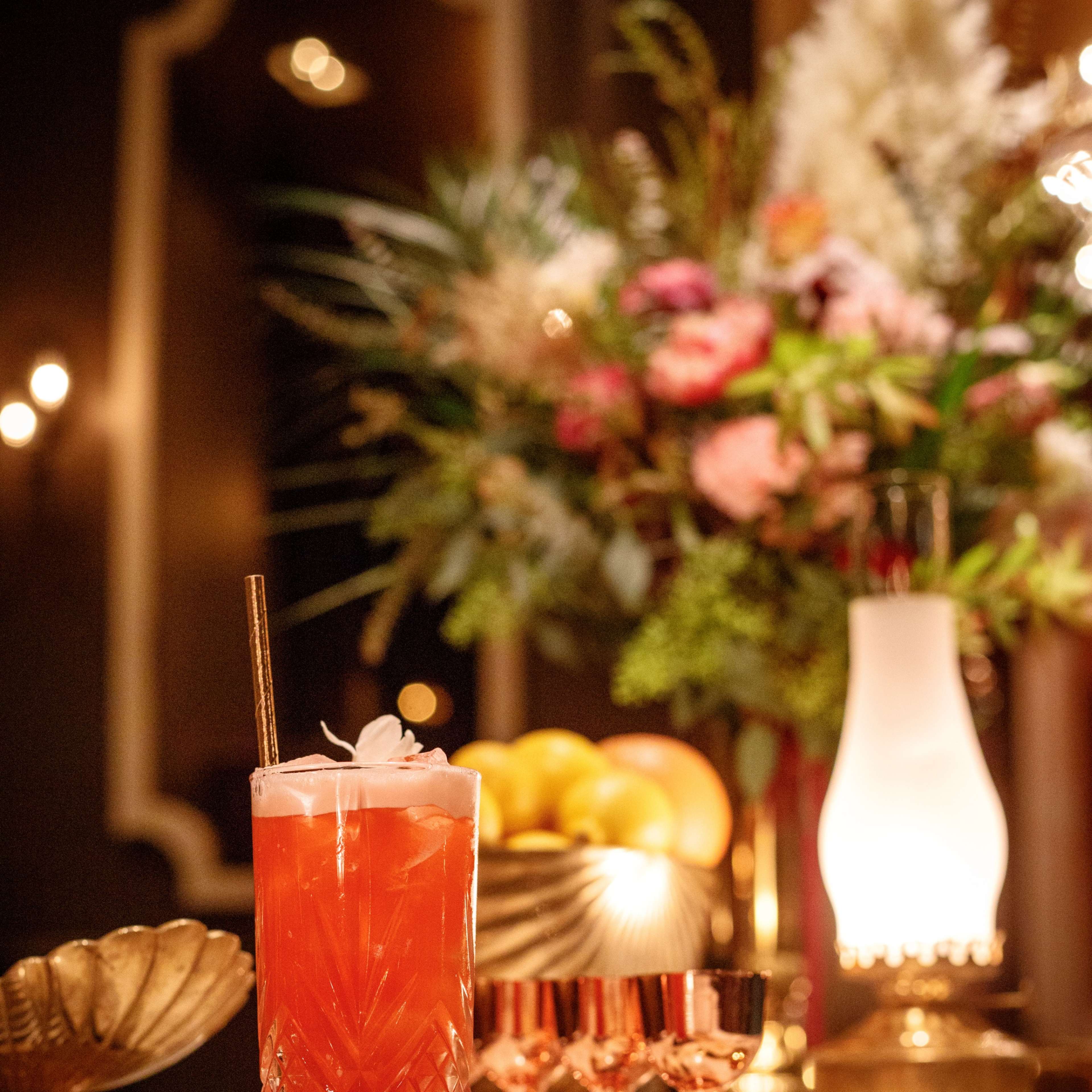 A tall glass of a vibrant cocktail sits on a marble table, accompanied by rose gold shot glasses and an ornate vase filled with flowers and fruit.