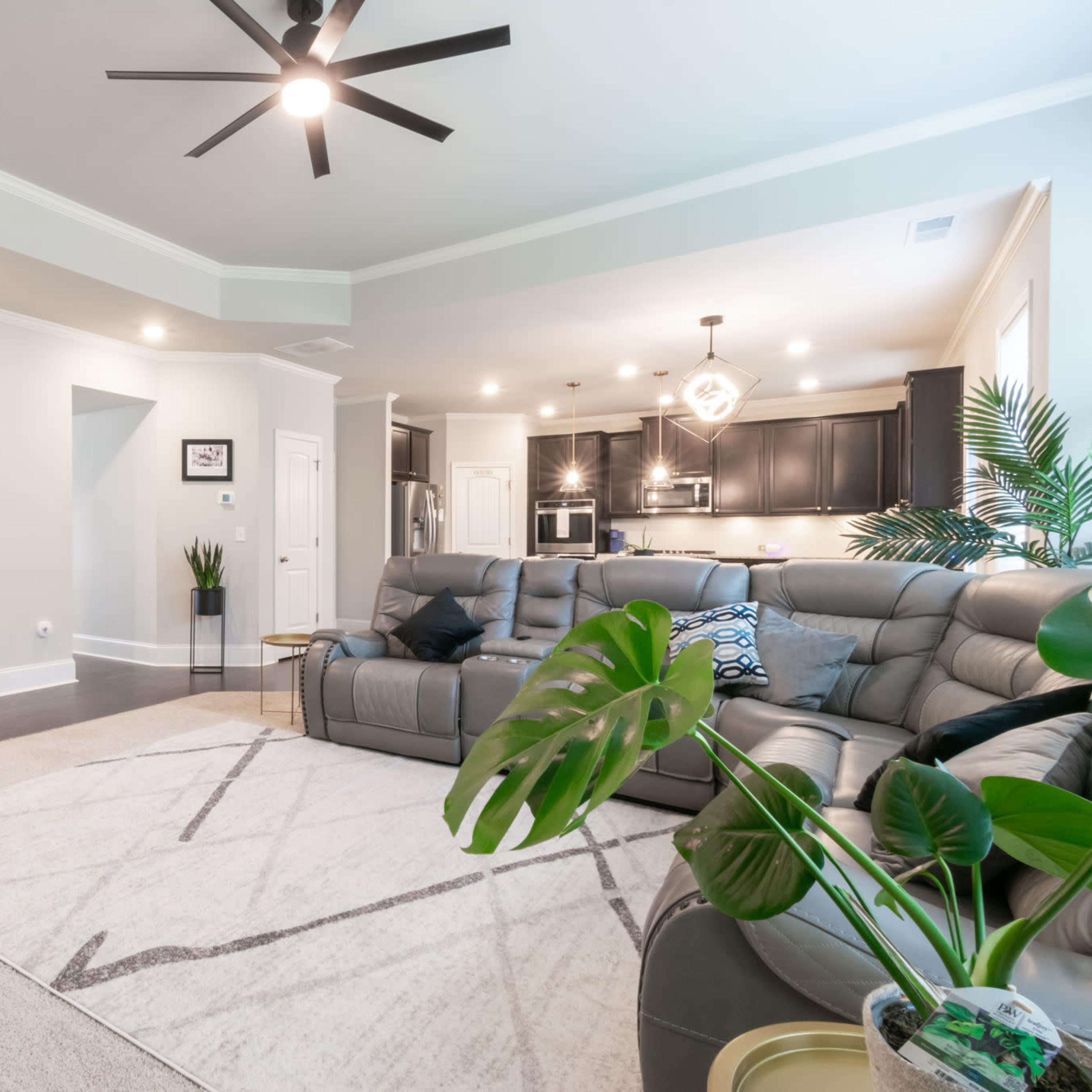 A spacious living room with a sectional sofa, a large rug, and a staircase leading to another level, along with modern kitchen appliances visible in the background.