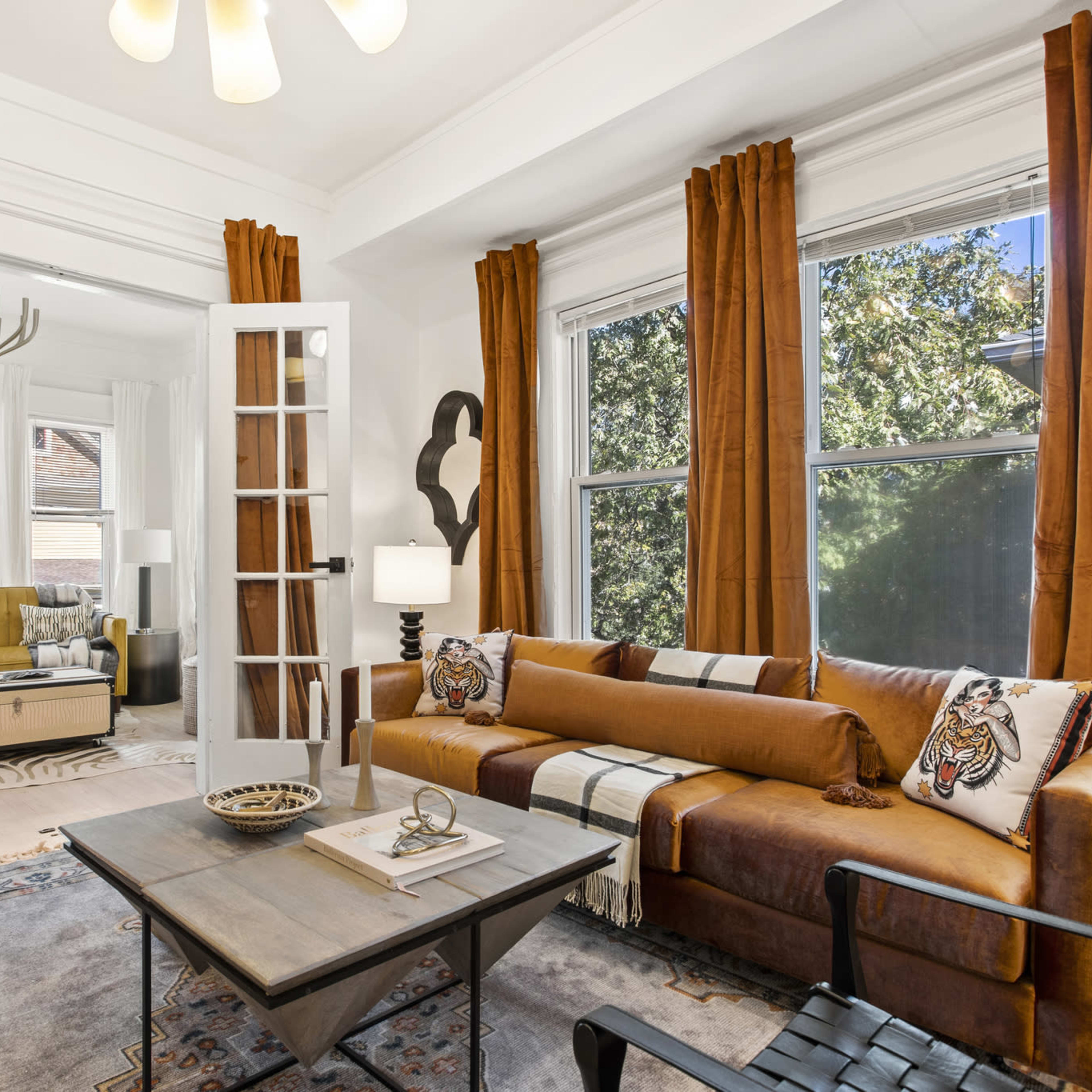 A cozy living room features a brown sofa, a coffee table, and large windows dressed with orange curtains, with a glimpse of a second room visible through French doors.