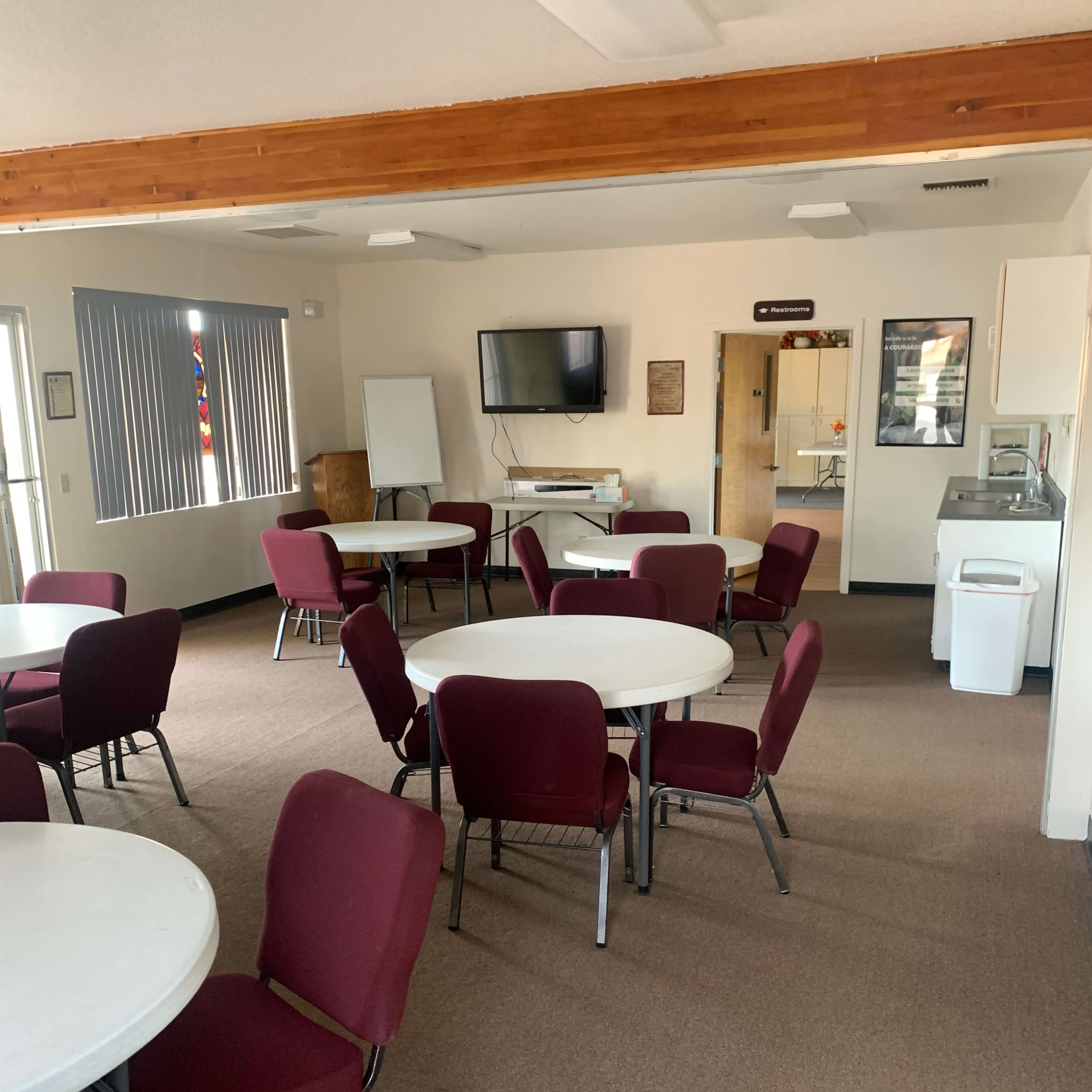 An empty room with several round tables and chairs arranged neatly, and a kitchenette area in the corner.