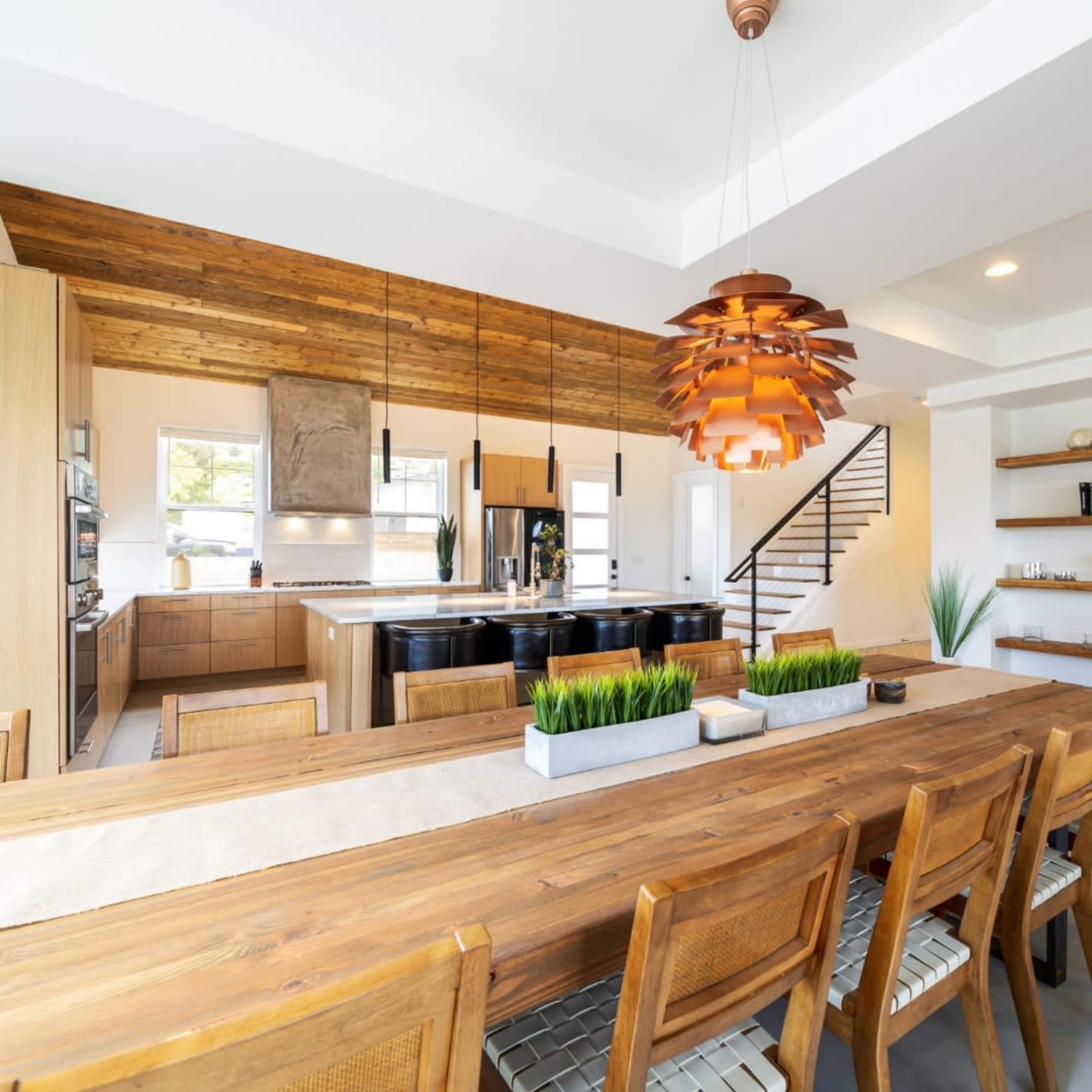 A spacious kitchen and dining area feature a large wooden table, modern lighting, and a mix of wooden and sleek cabinetry.