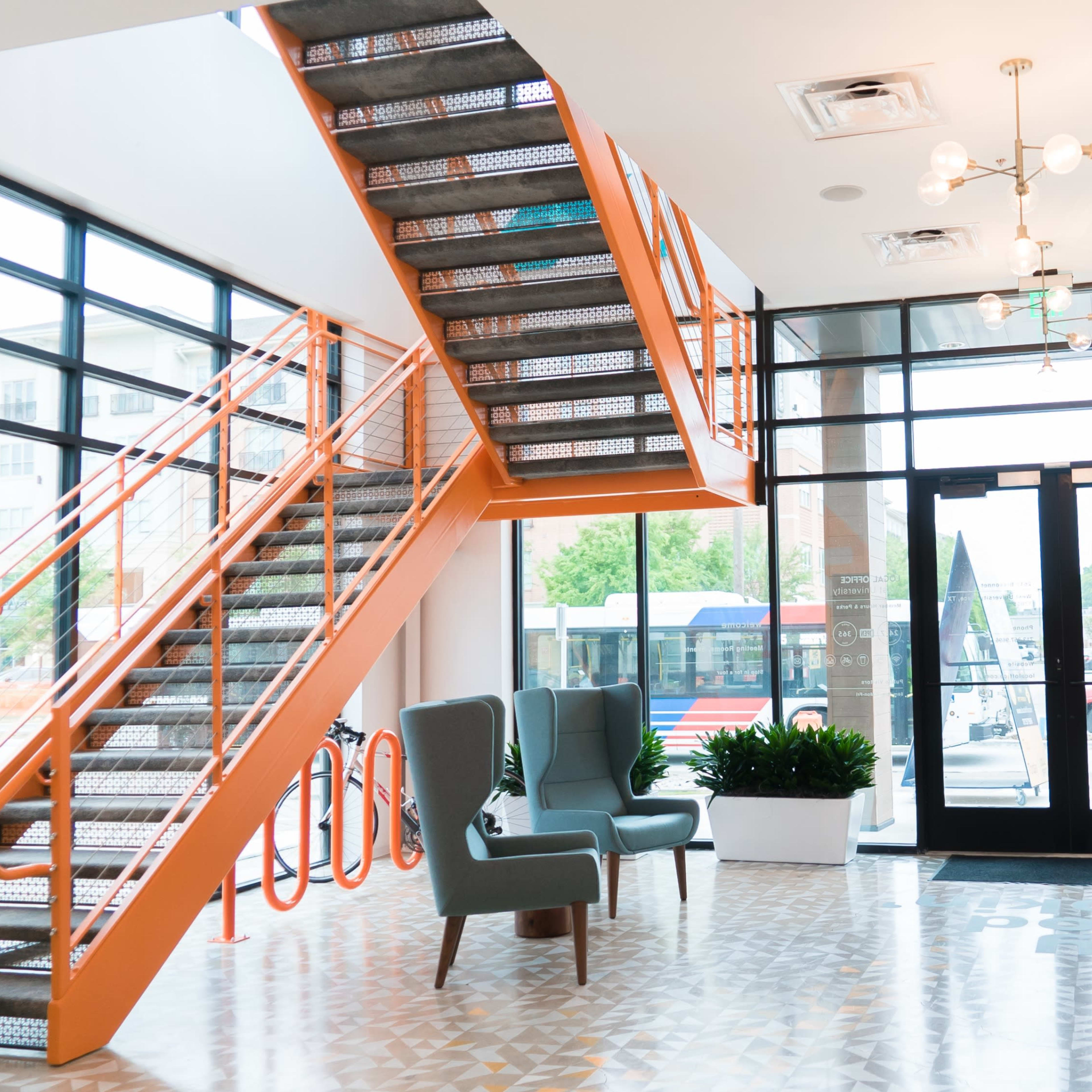 A modern lobby area featuring a staircase with an orange railing, two green chairs, and a row of mailboxes against a large window.