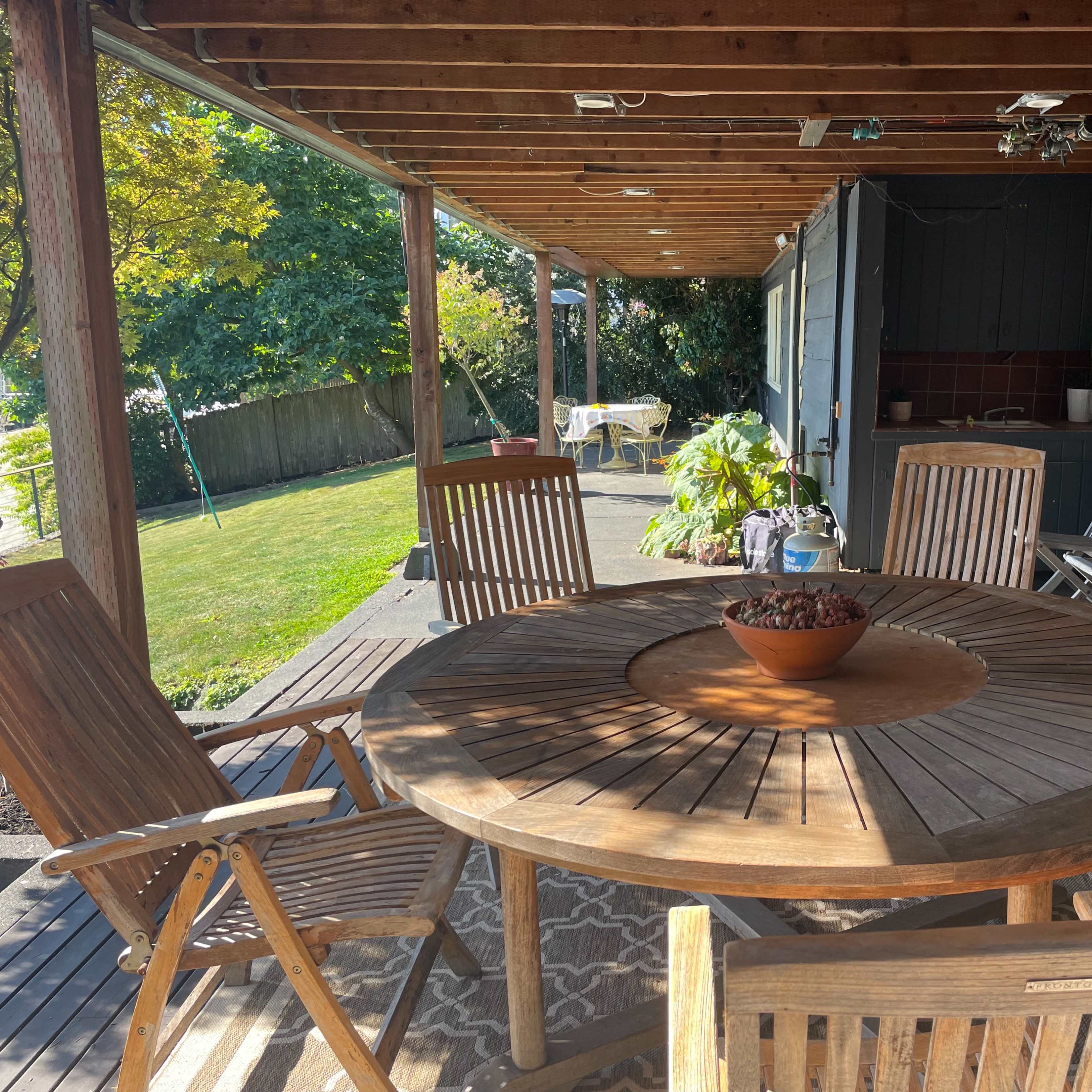 A wooden patio with a round table and several chairs sits under a covered area, surrounded by greenery.