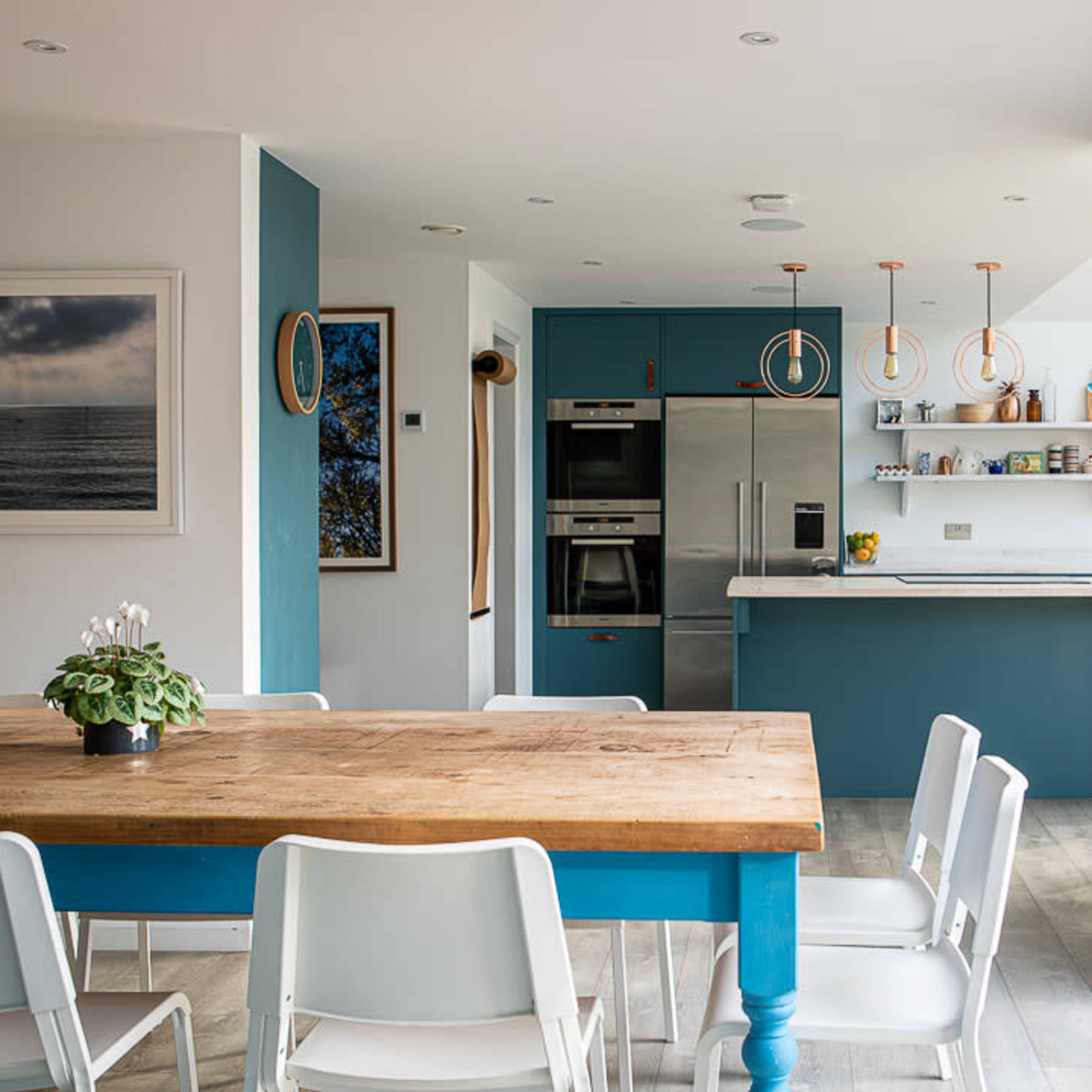 A modern kitchen and dining area featuring a wooden table surrounded by white chairs, with teal cabinetry and appliances in a well-lit space.