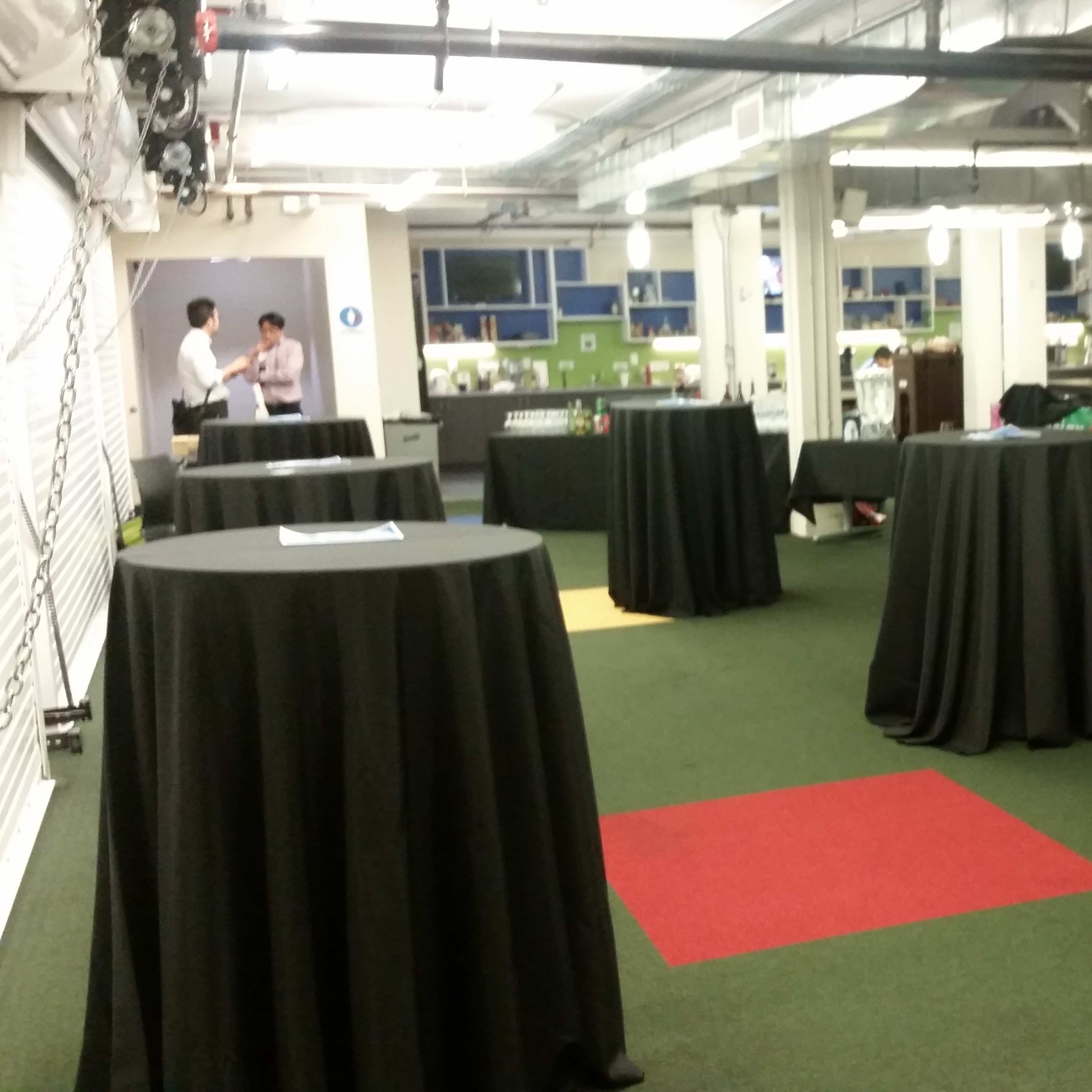 A large, open space with several tall, round tables covered in black tablecloths, surrounded by a green carpet and colored floor sections.