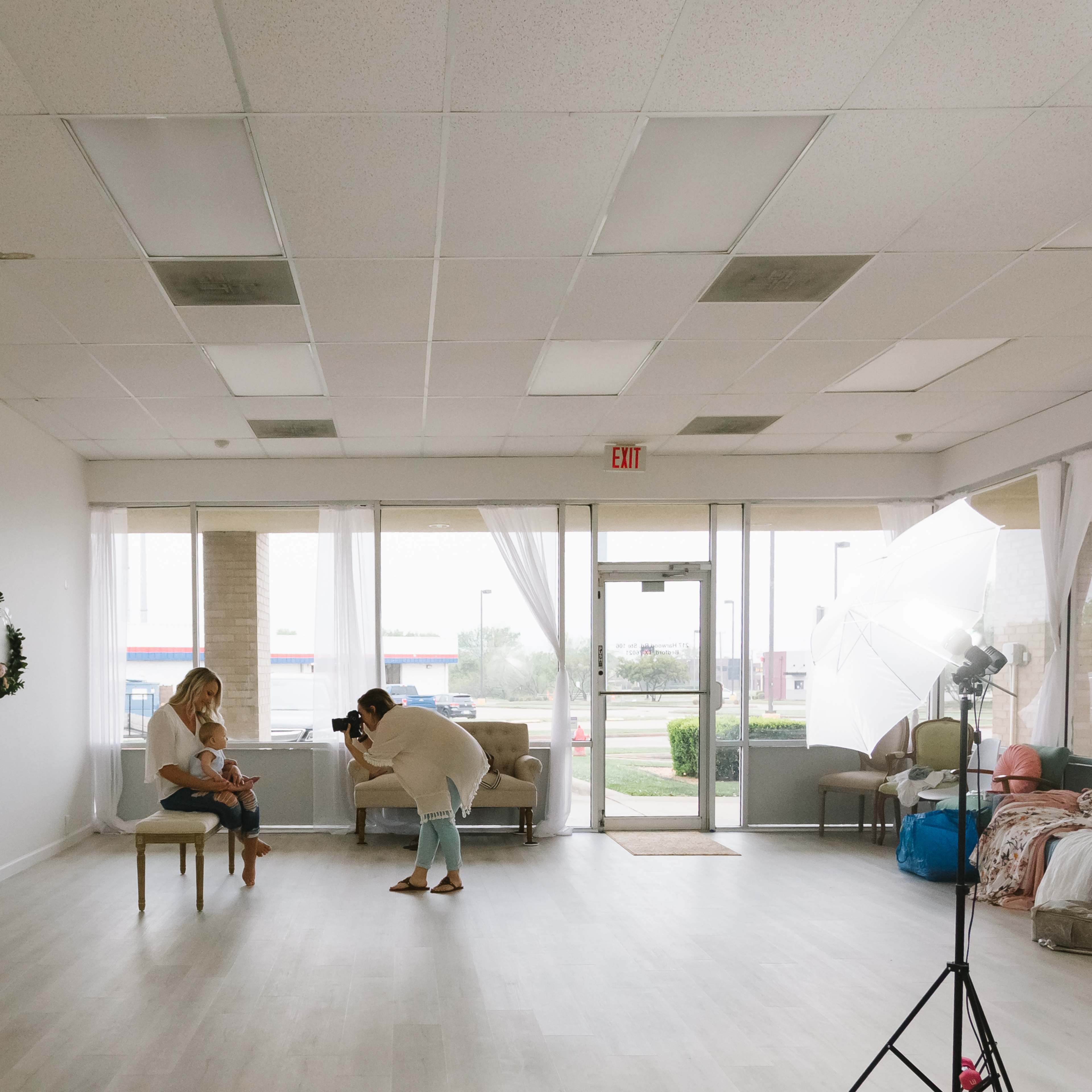 A photographer is capturing images of a child sitting on a chair in a spacious, well-lit studio with large windows.