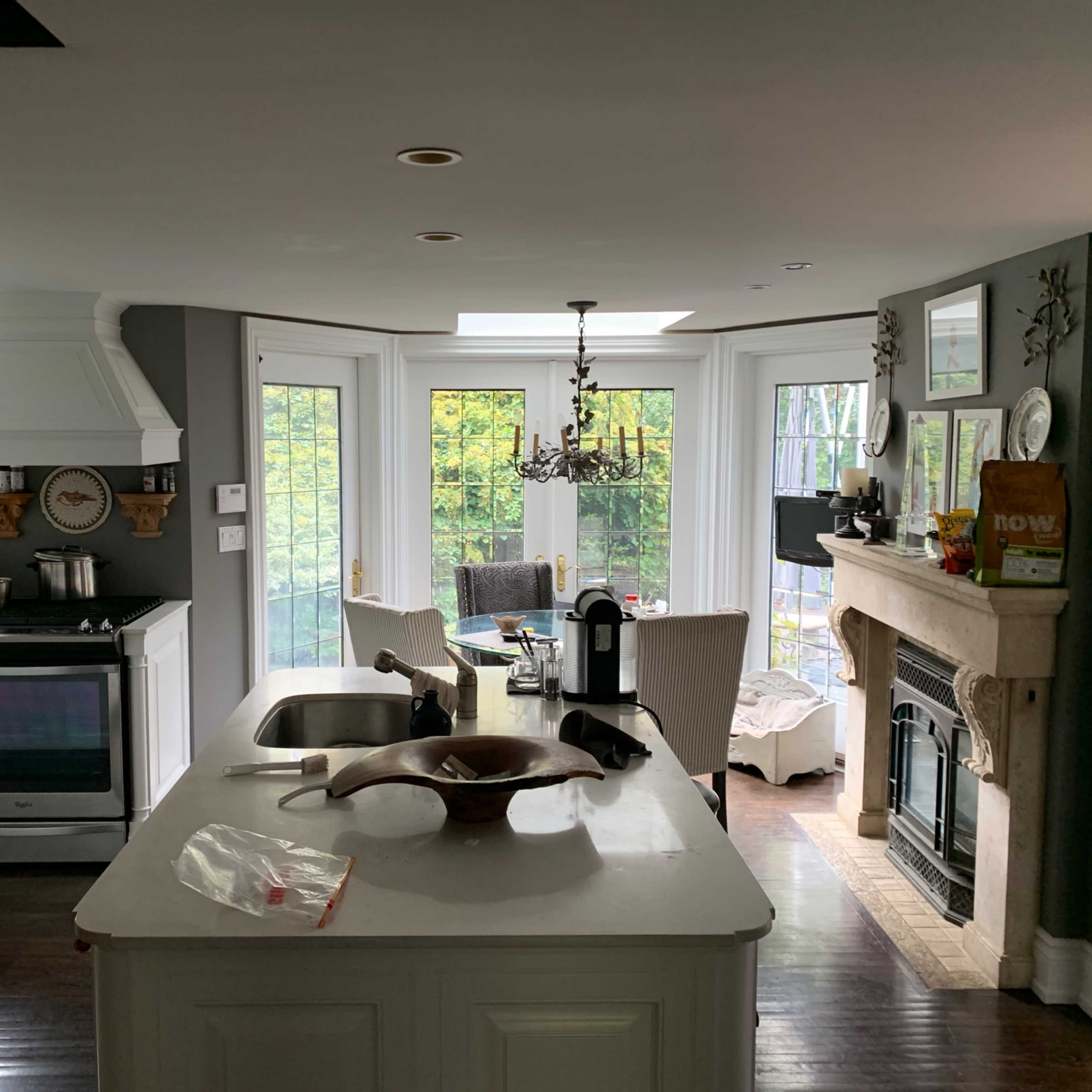 A modern kitchen with white cabinetry and a central island, featuring a view of a dining area with a fireplace and large windows.