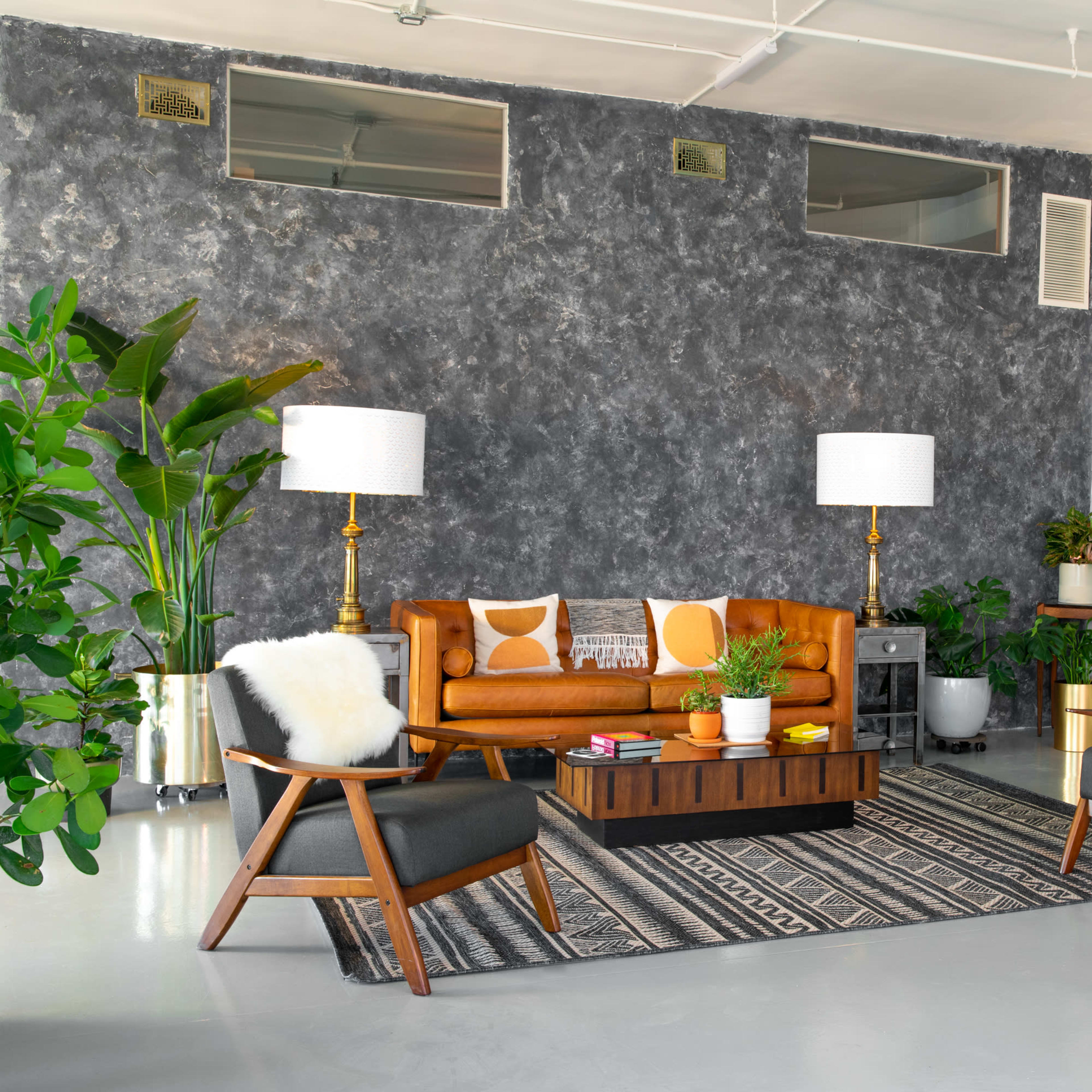 A modern living room with a brown leather sofa, two gray armchairs, a wooden coffee table, and various indoor plants against a textured gray wall.