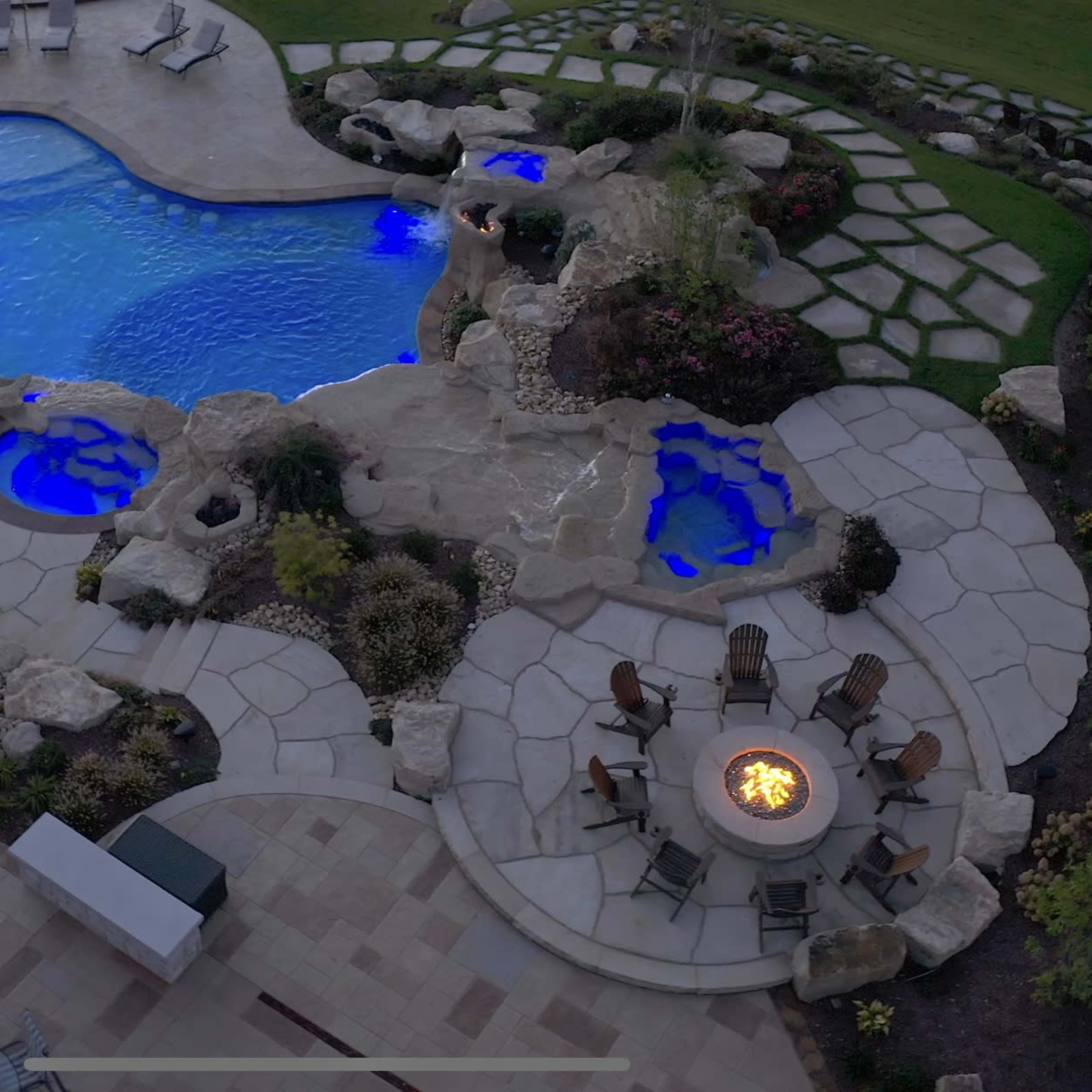 An aerial view shows a landscaped pool area with a blue swimming pool, stone pathways, and a fire pit surrounded by chairs.