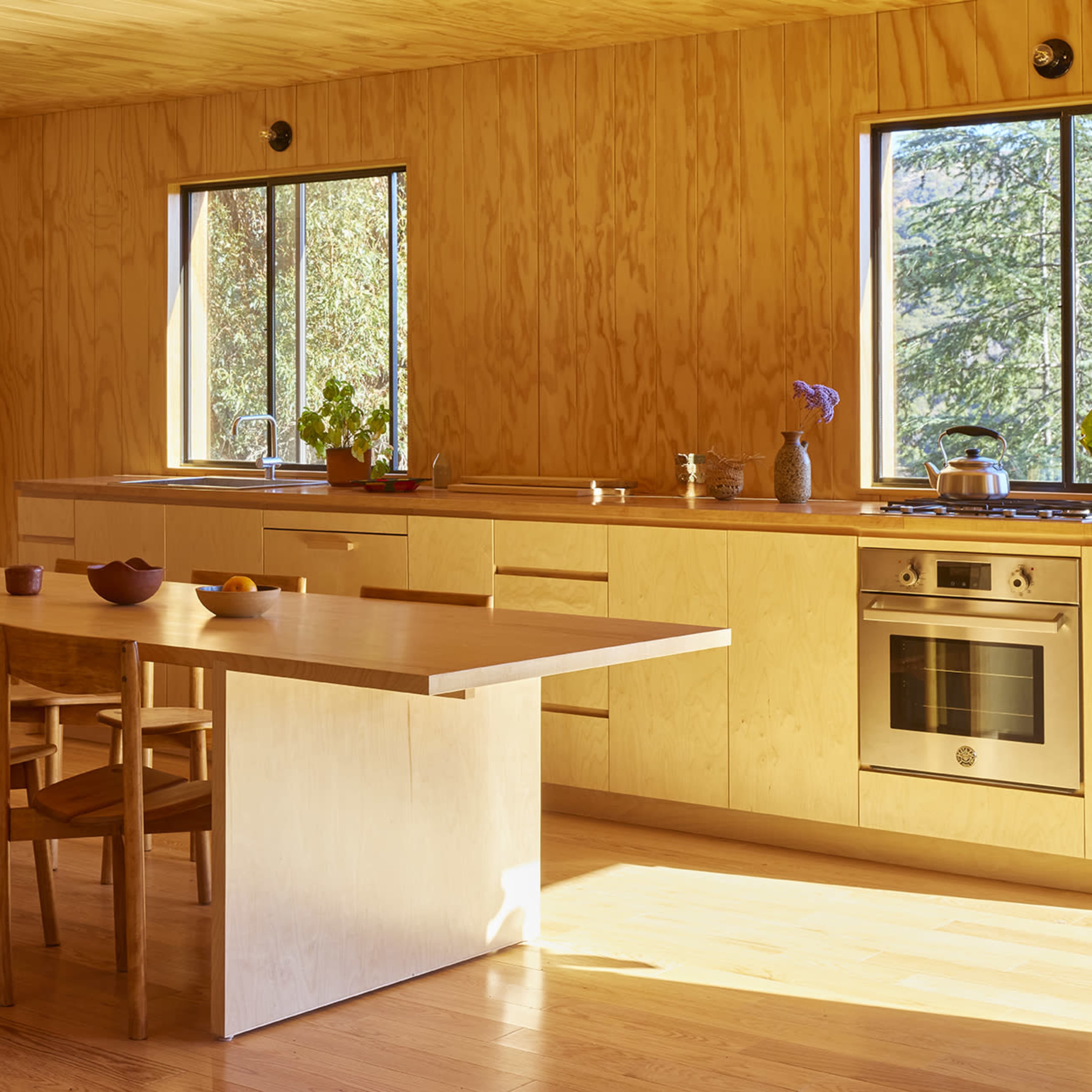 A modern kitchen with wooden cabinetry, a large table, and four chairs, illuminated by natural light from two windows.
