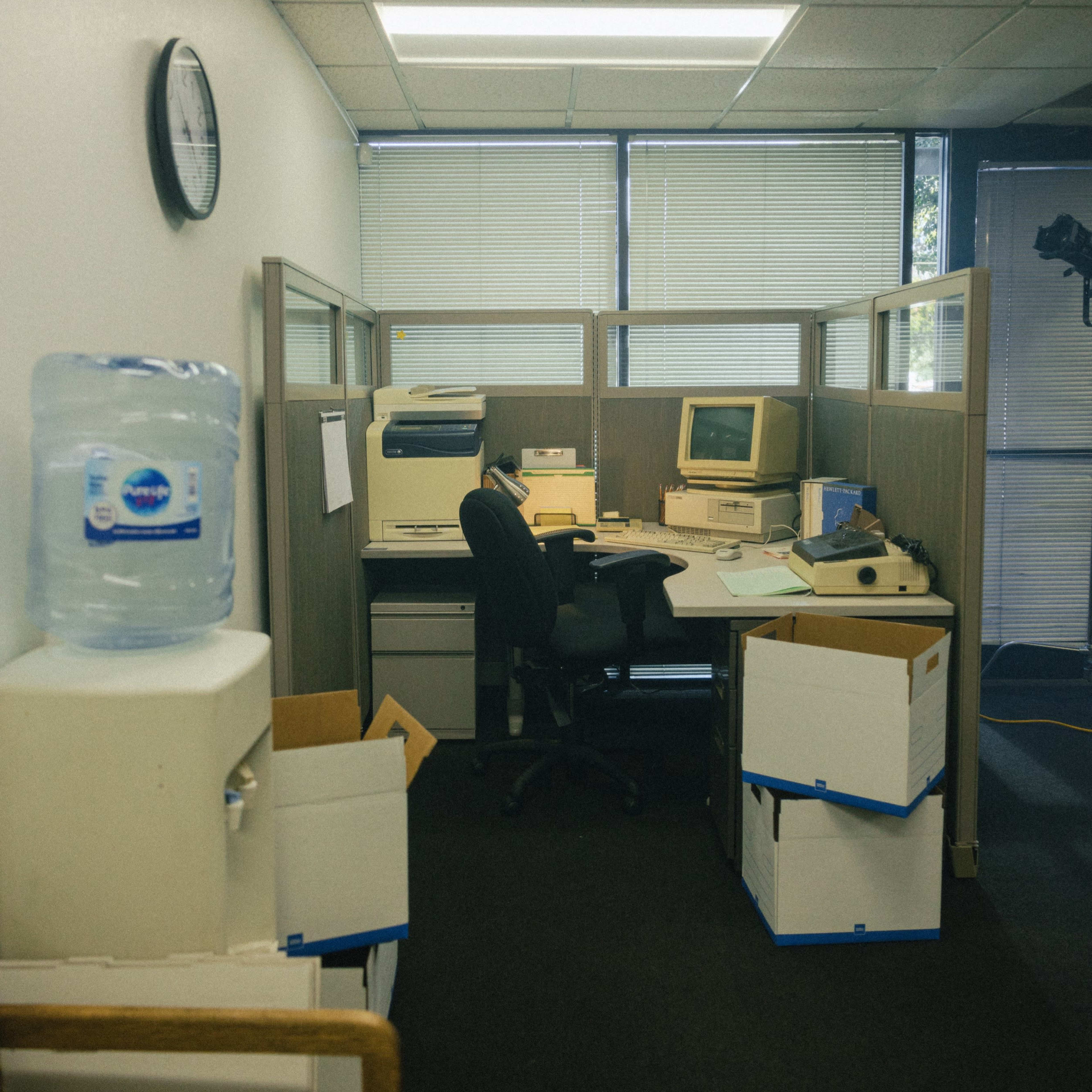 A small office cubicle with an old computer, a printer, boxes, and a water cooler in a dimly lit workspace.