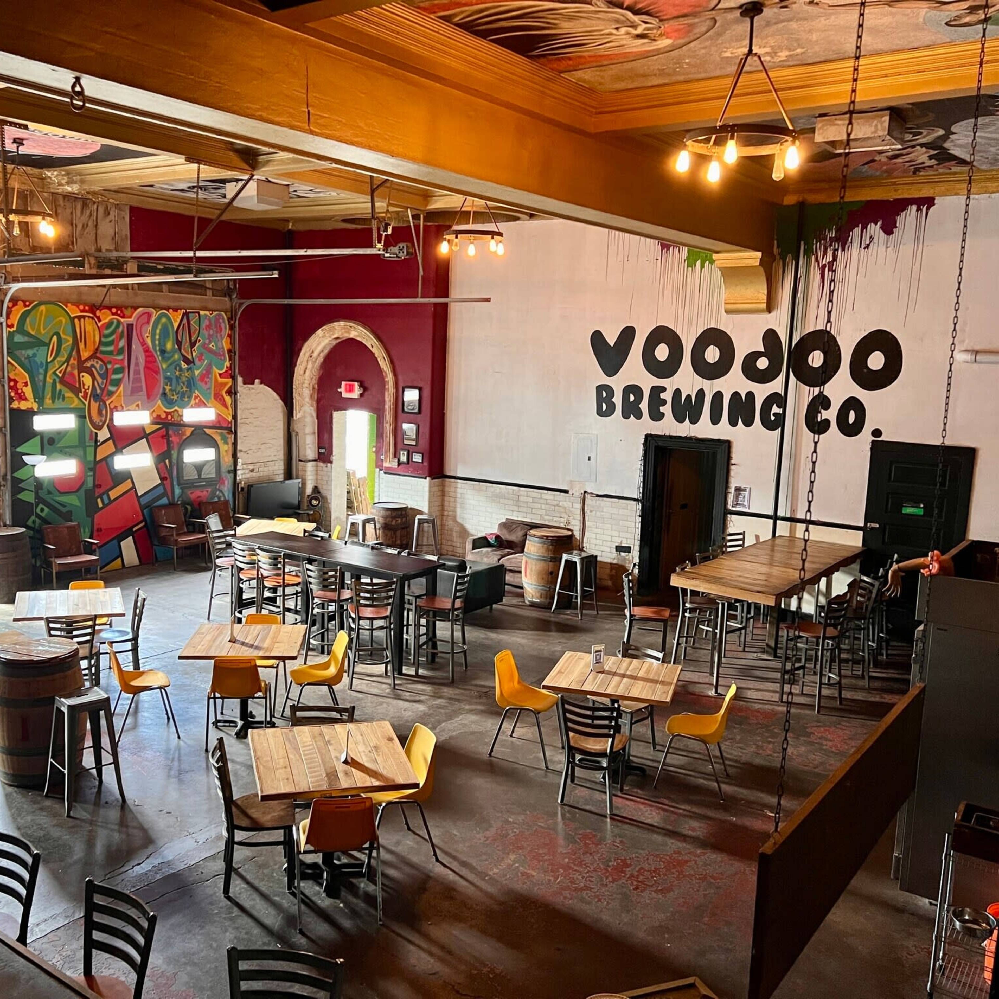 The interior of Voodoo Brewing Co. features an open seating area with wooden tables, yellow chairs, and colorful wall murals.
