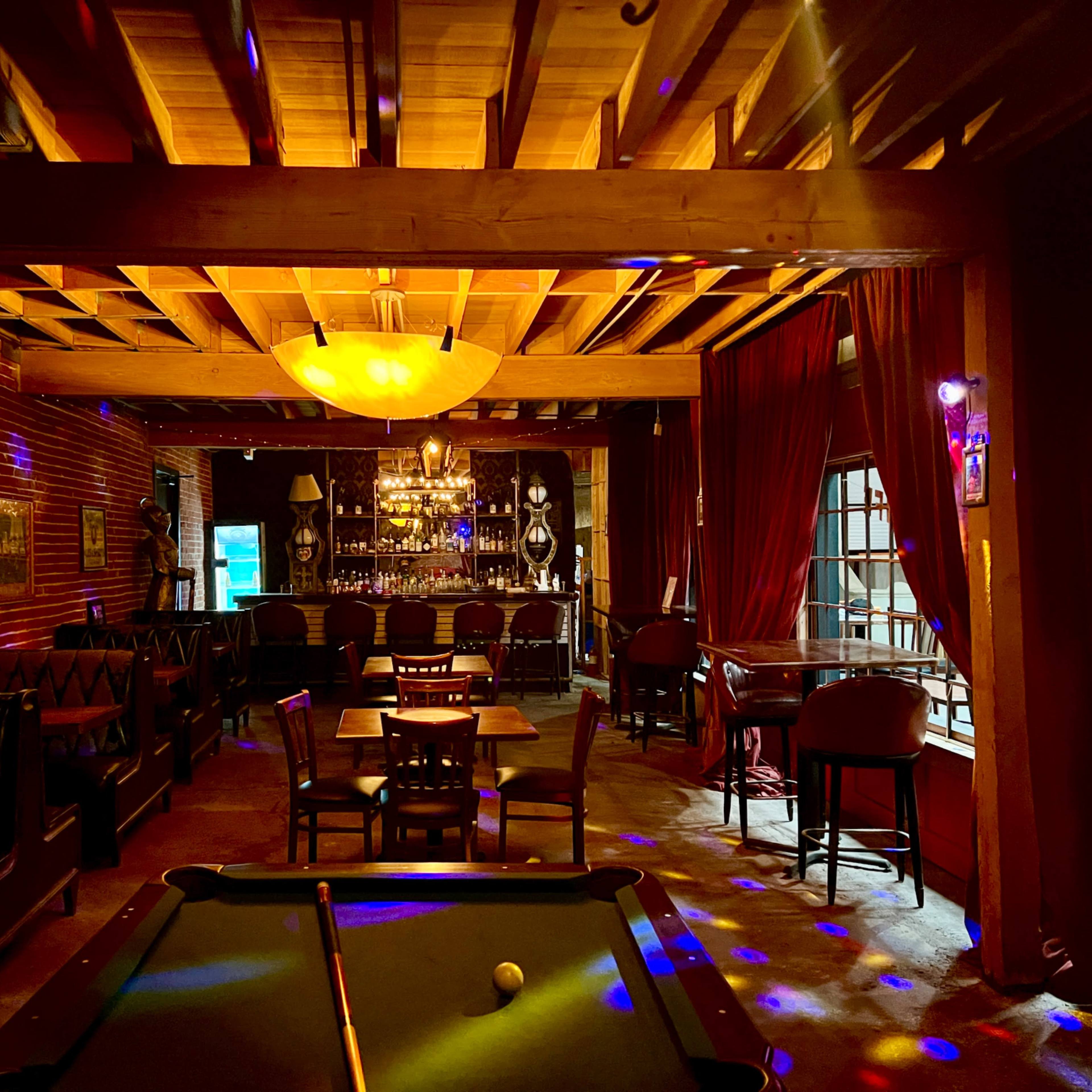 A dimly lit bar with a pool table in the foreground, surrounded by wooden furnishings and brick walls, and soft lighting from a ceiling fixture.
