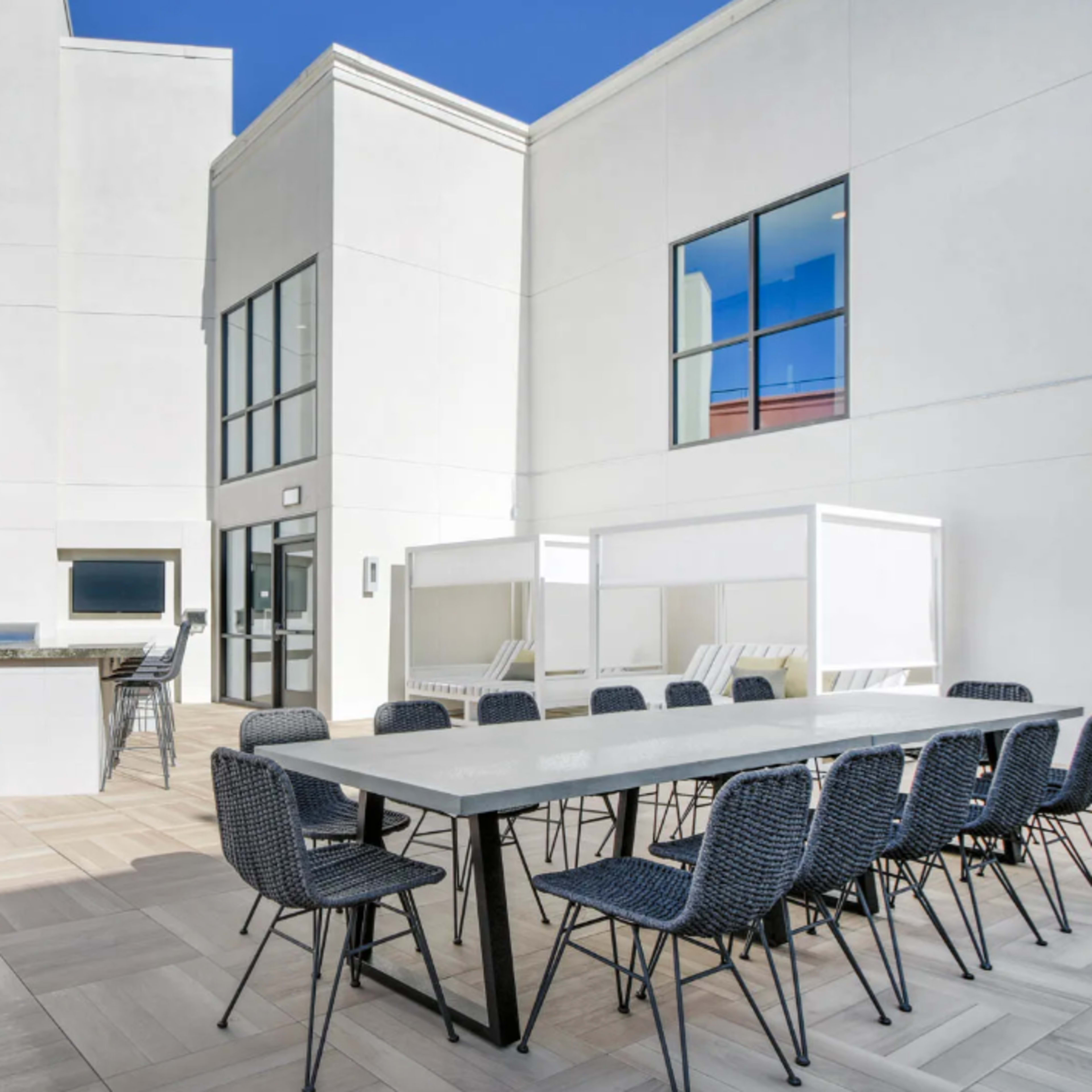 A spacious outdoor patio area with a large dining table surrounded by chairs, a grill station, and white cabanas against a backdrop of modern white buildings.