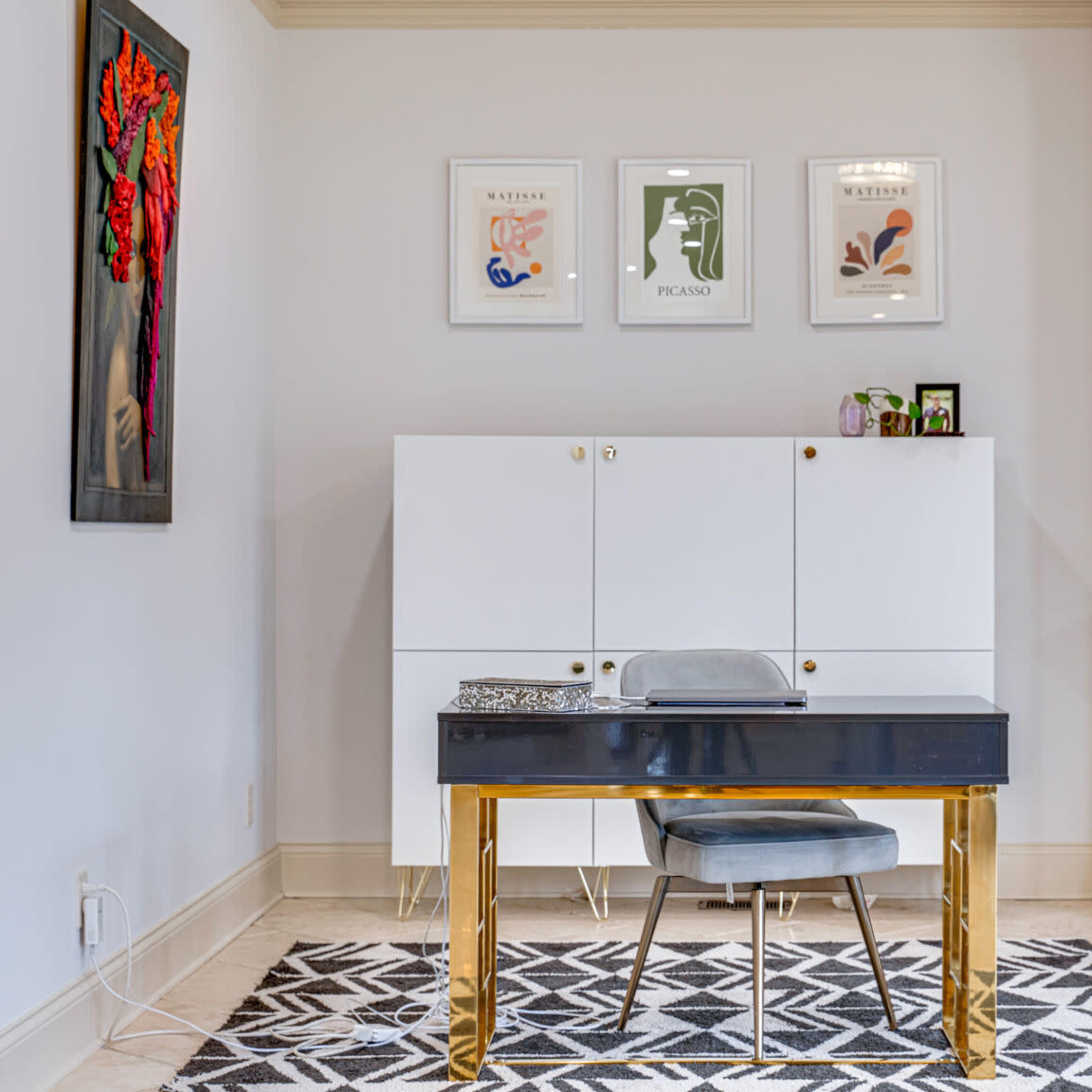 A modern home office with a desk, a chair, framed artworks on the wall, and a fireplace.
