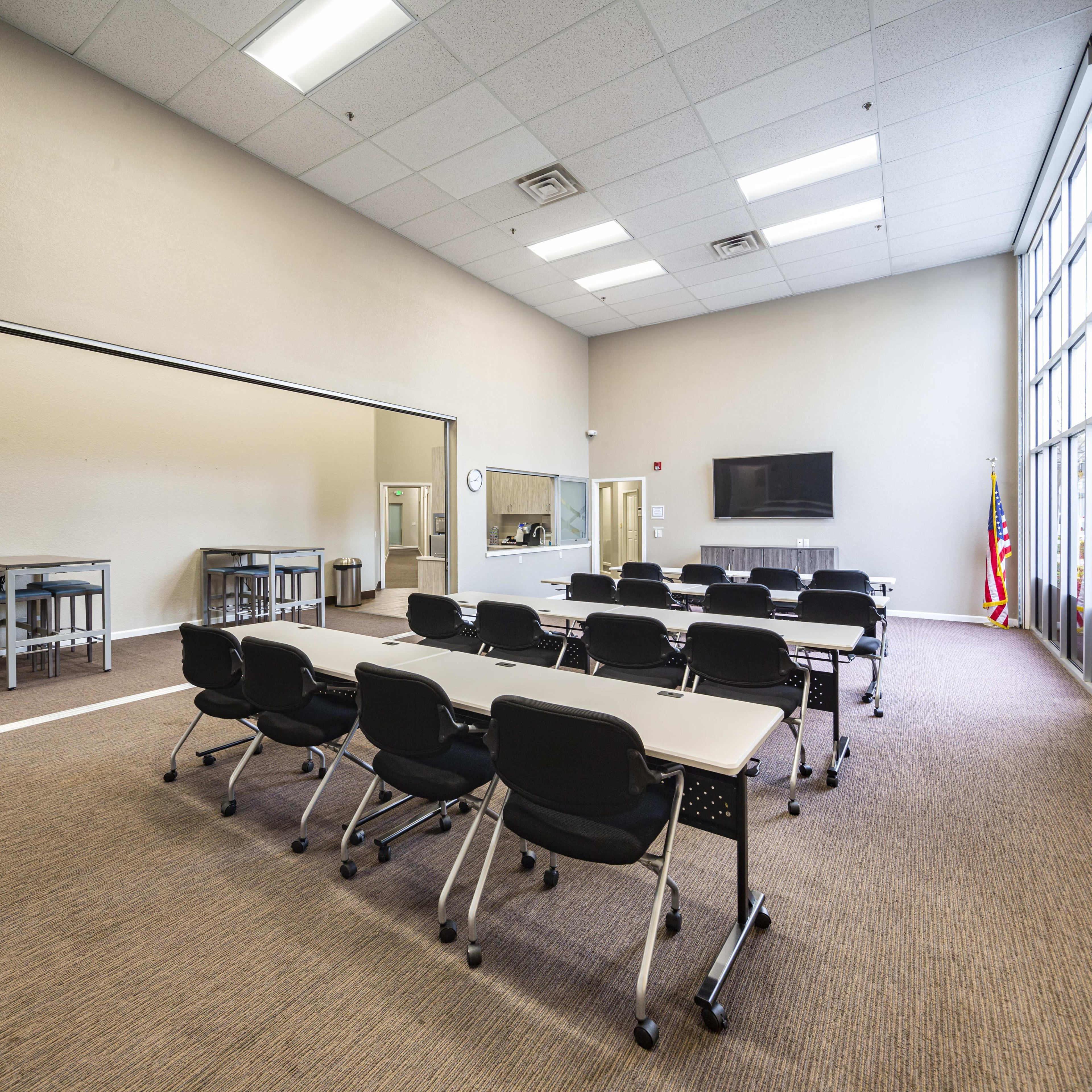 A spacious, well-lit classroom with multiple tables and chairs arranged in a grid, featuring a large wall-mounted television and an American flag in the corner.