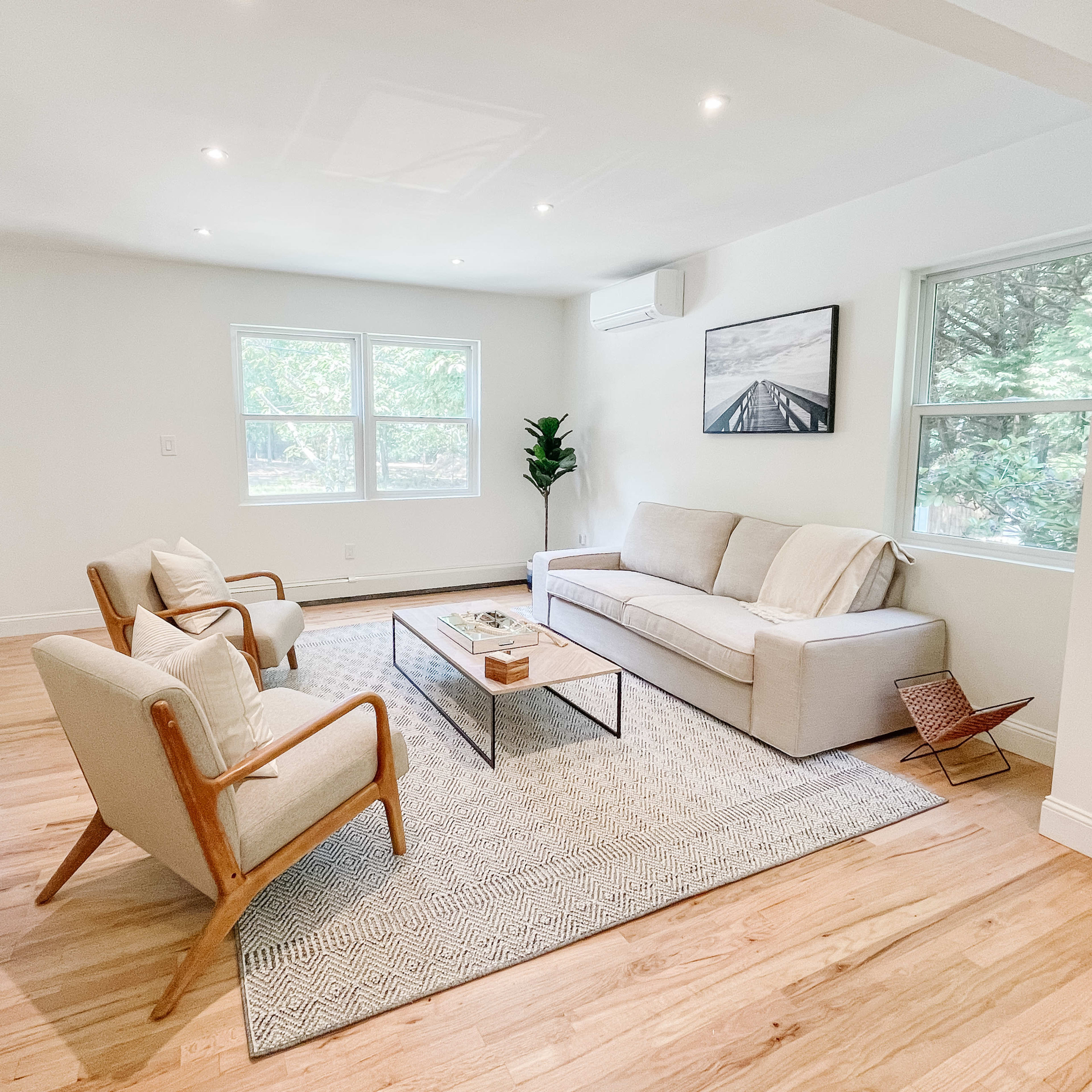 The living room features a light-colored sofa, two wooden armchairs, a coffee table, and large windows that provide natural light.