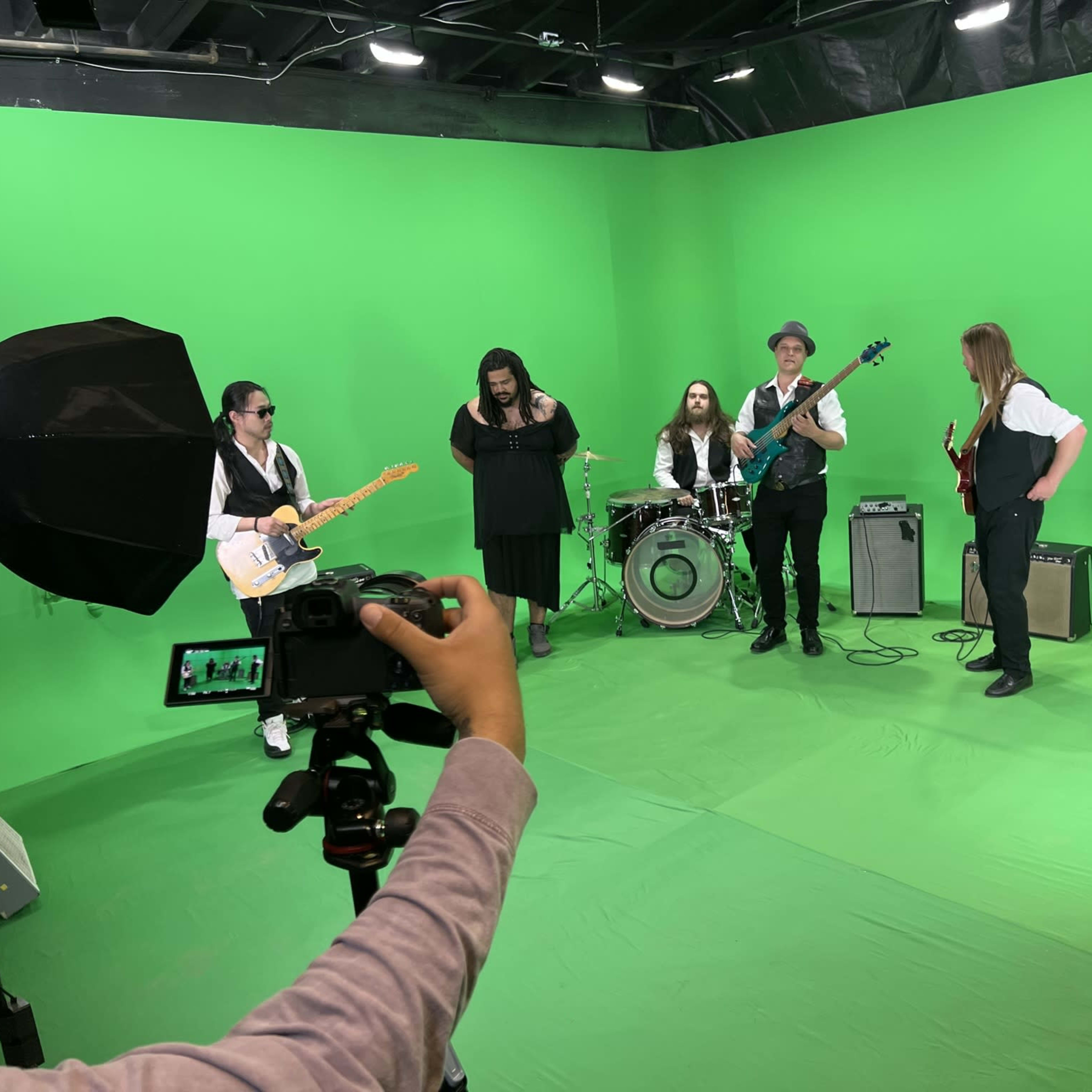 A band is performing in front of a green screen, with instruments set up and a camera focusing on one of the musicians.