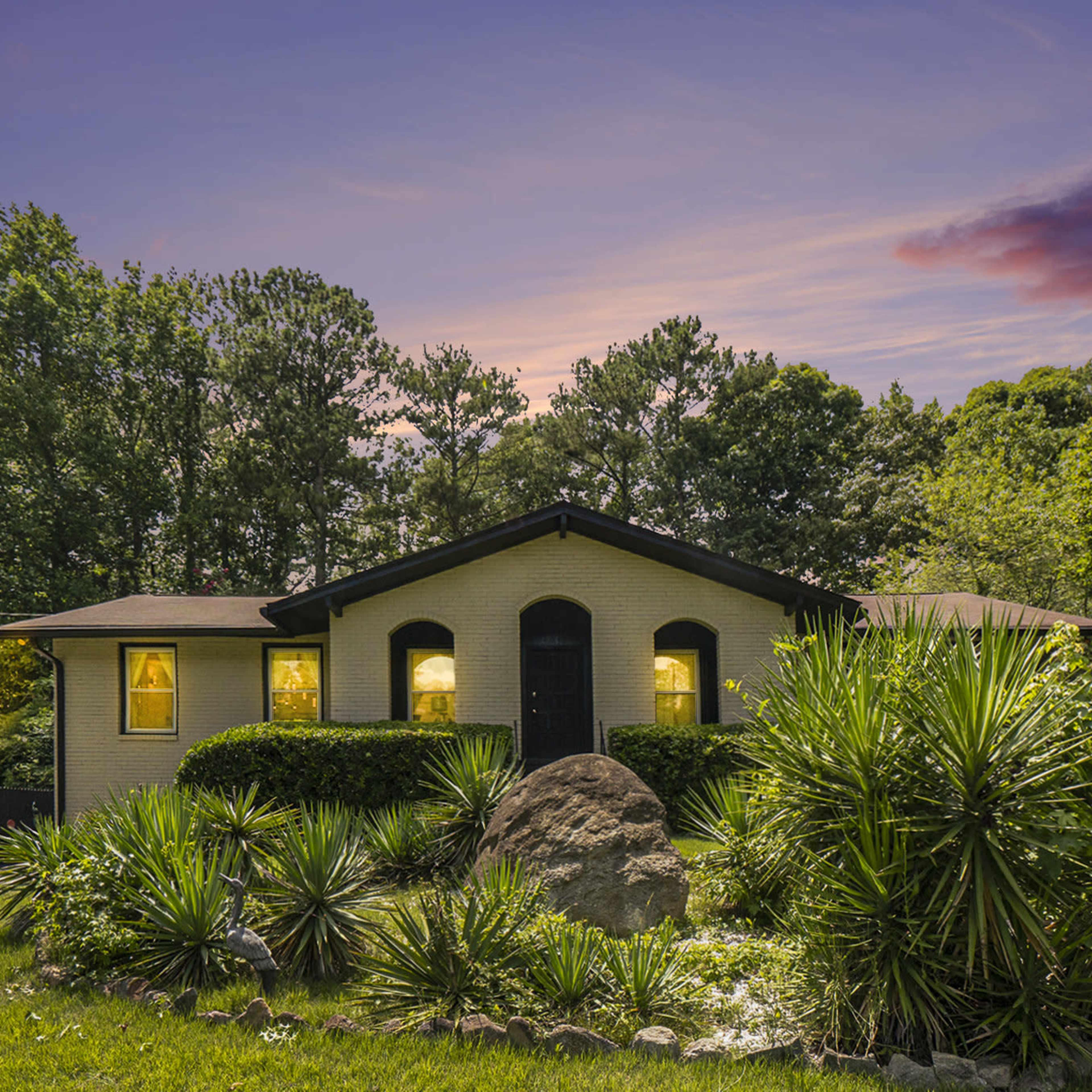 A single-story house with illuminated windows is set among landscaped greenery and trees at dusk.