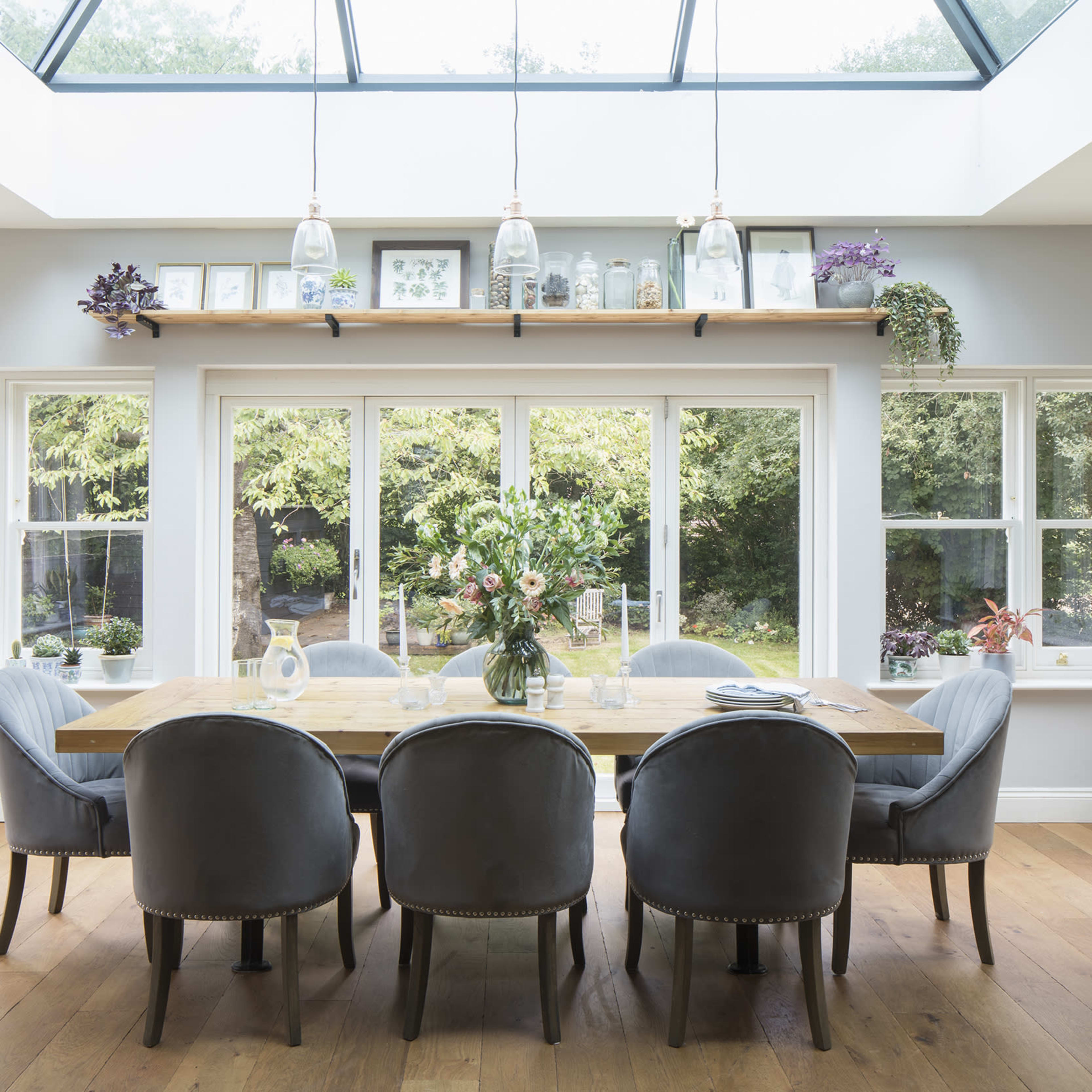 The dining room features a long wooden table surrounded by upholstered chairs, with large windows providing a view of the garden outside.