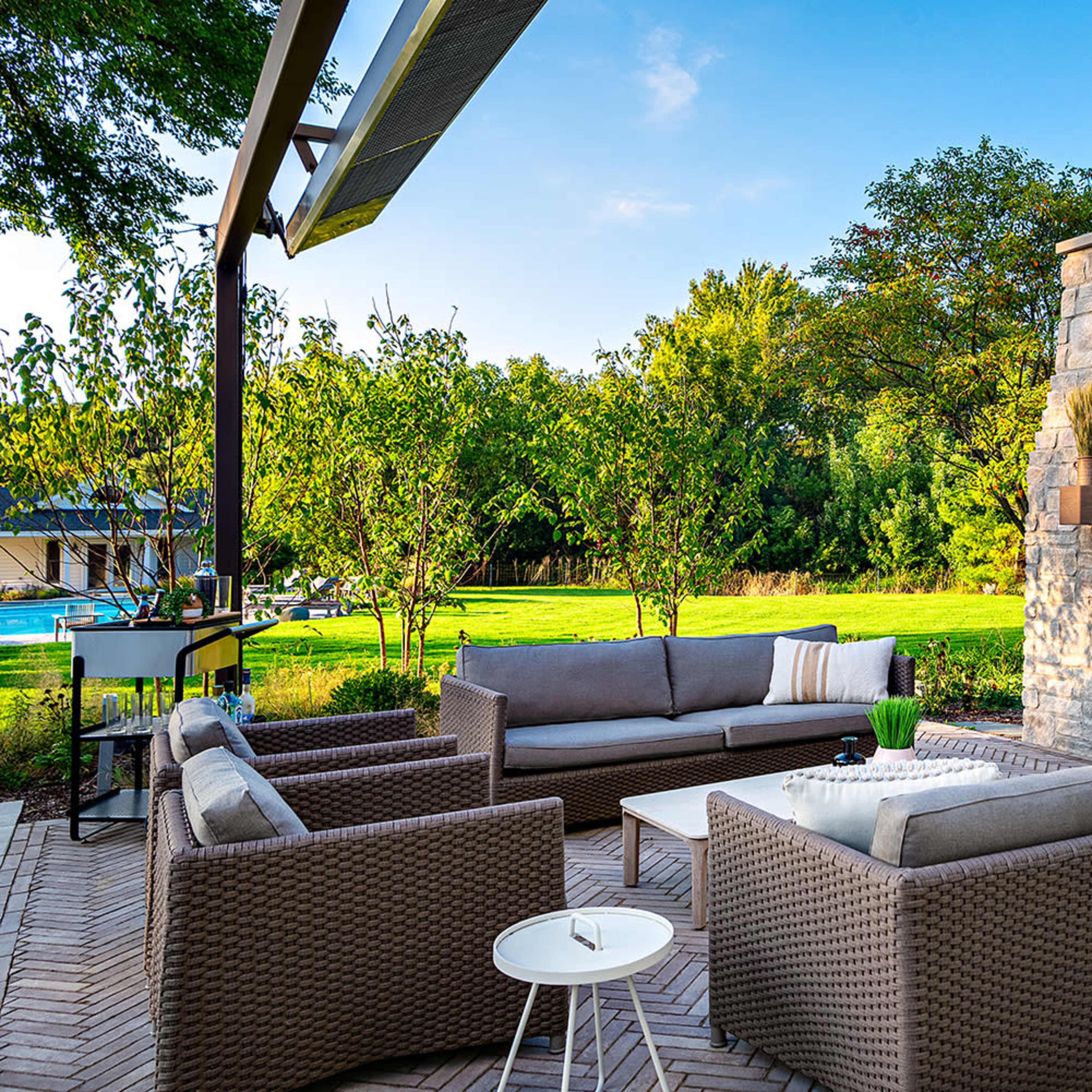 A patio with a seating area featuring wicker furniture and a mounted TV above a stone fireplace, surrounded by greenery and a view of a pool.