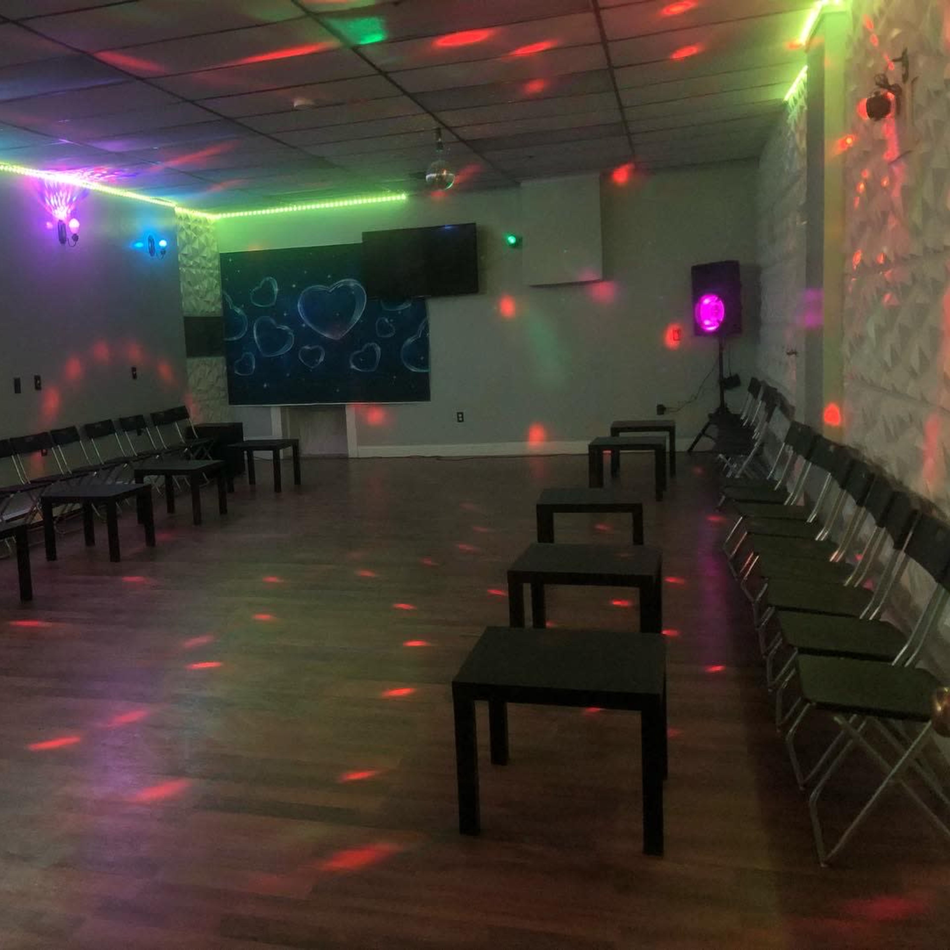 A spacious room features rows of chairs and tables, illuminated by colorful lights reflecting on the walls.