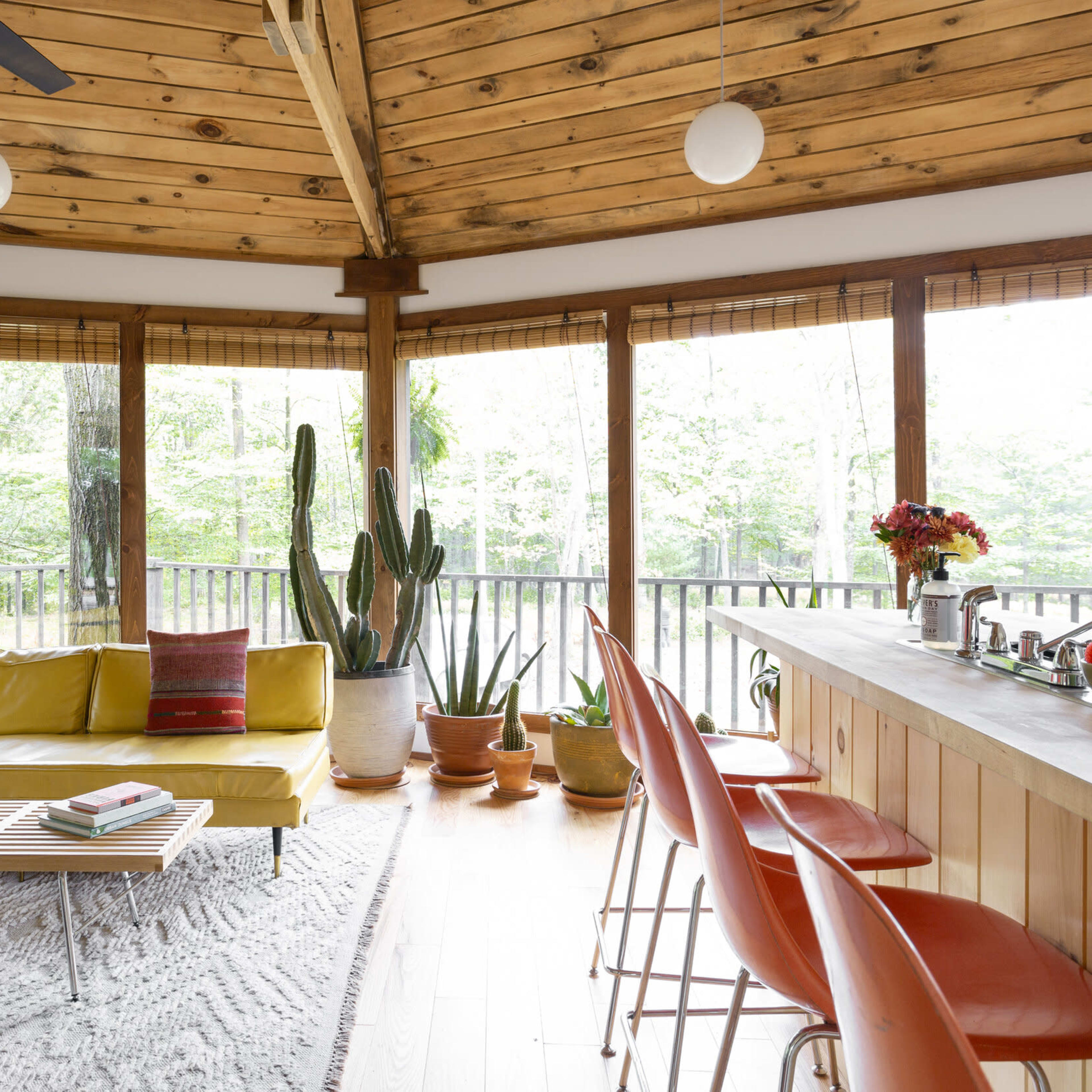 A modern, circular living space features a yellow sofa, a wooden ceiling, a bar with colorful stools, and large windows overlooking a forested area.