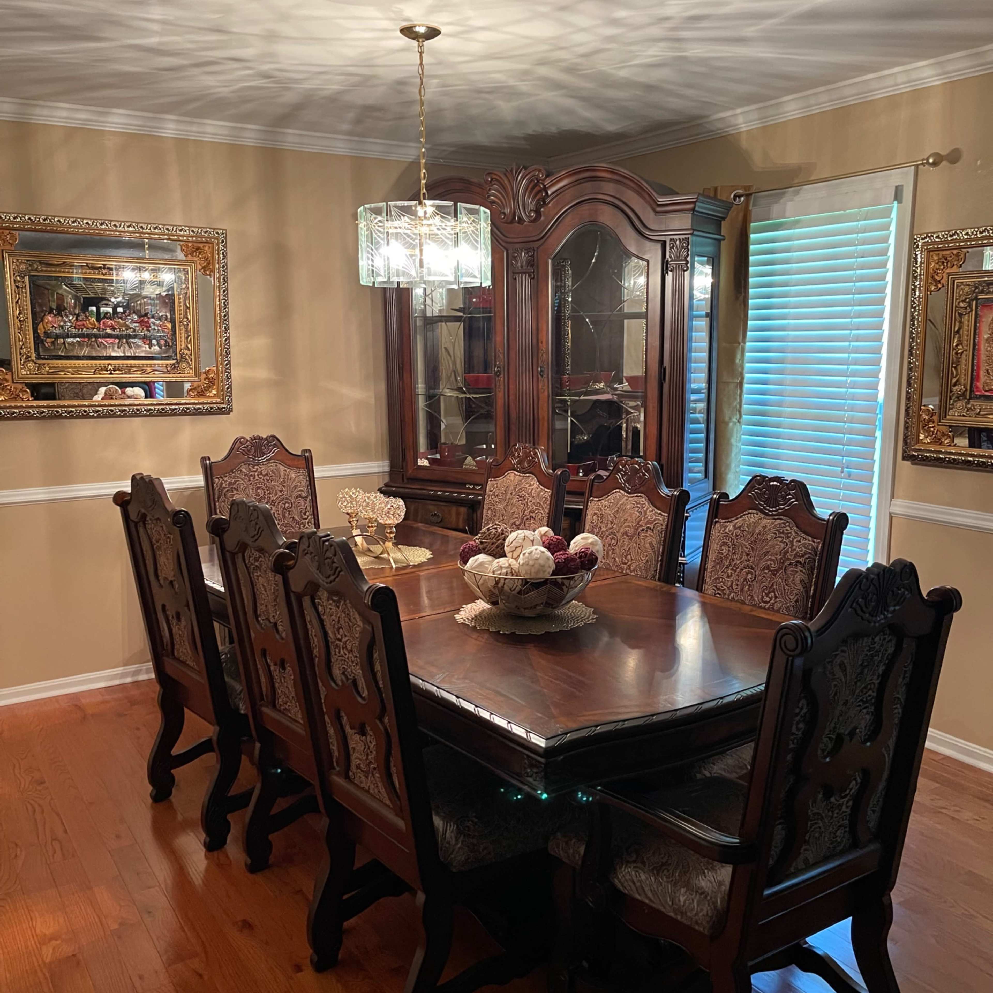 A wooden dining table with eight ornate chairs is centered in a warmly lit dining room, surrounded by elegant decor and a china cabinet.