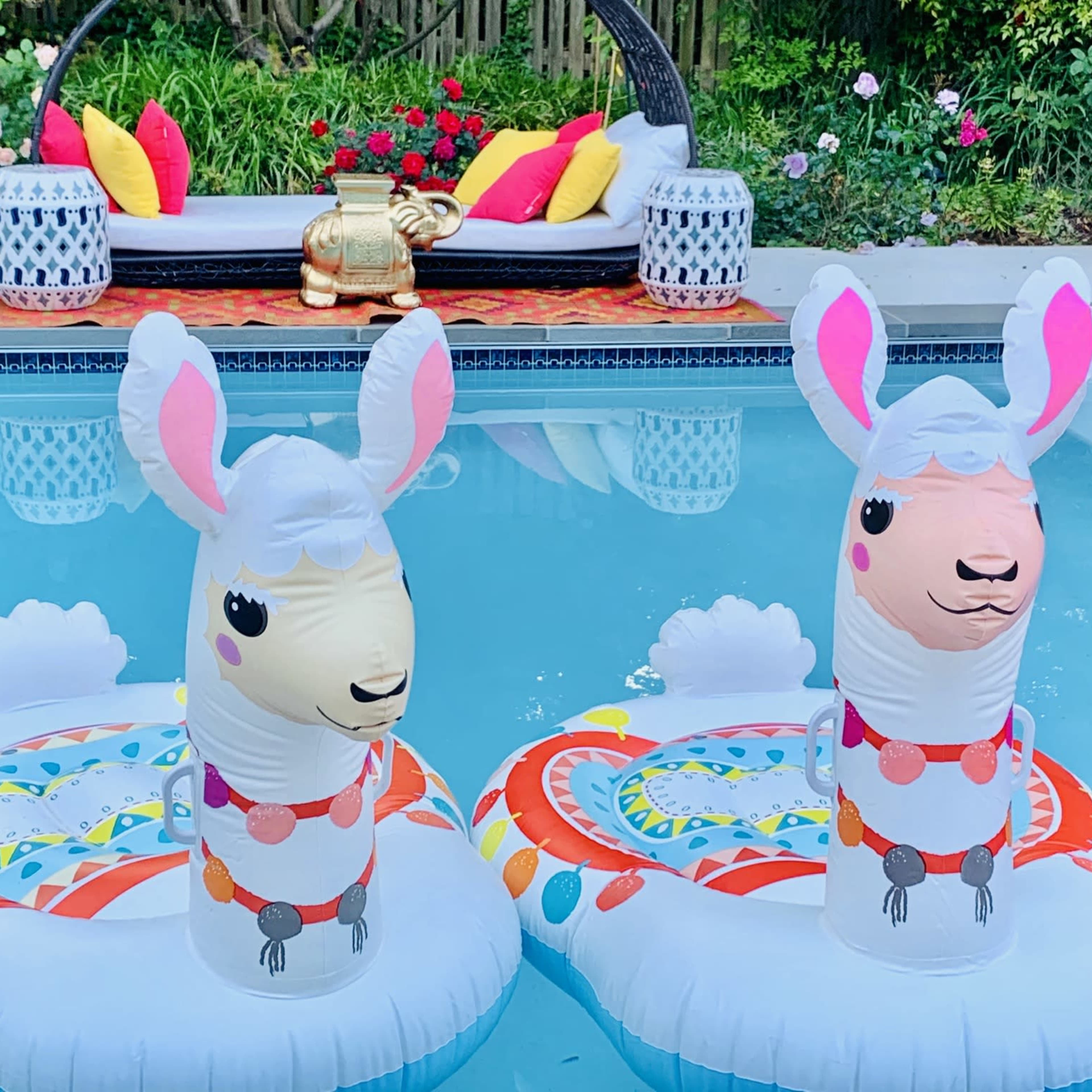 Two inflatable llama pool floats are positioned in a swimming pool, surrounded by a landscaped yard featuring colorful flowers and a lounge area in the background.